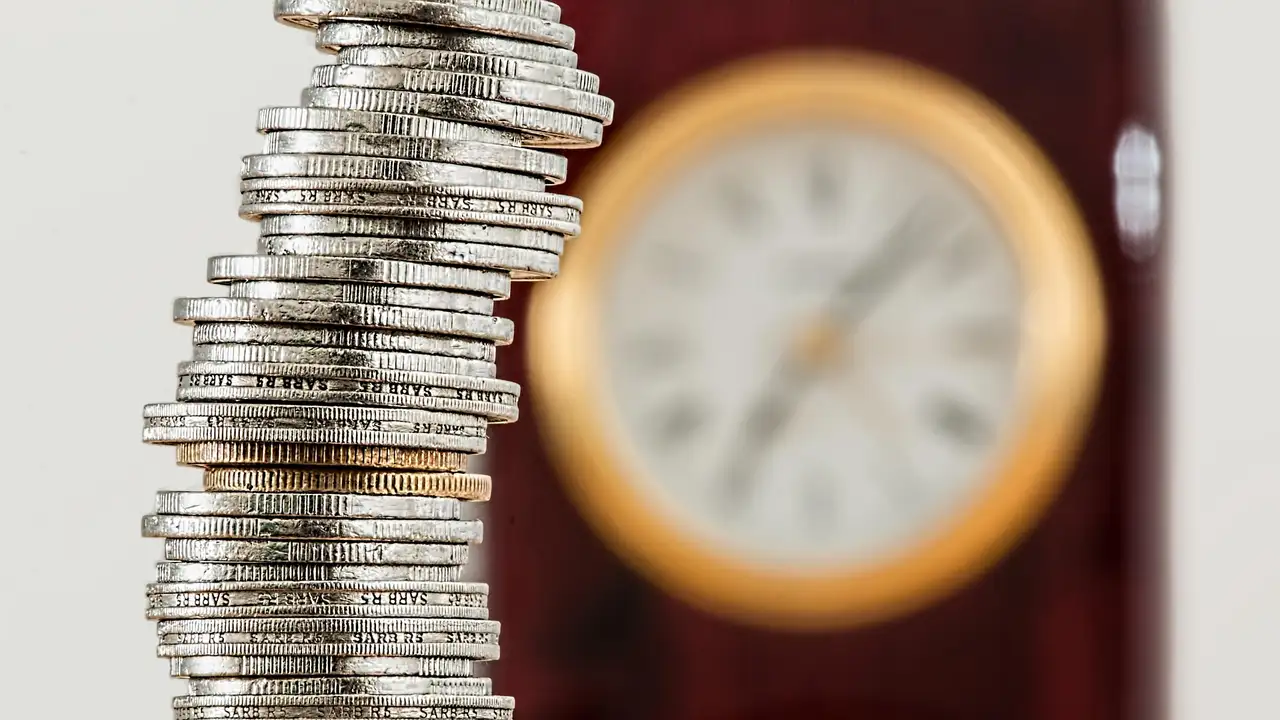 Imagem mostra pilha de moedas e relógio ao fundo