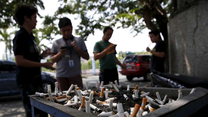 Men smoke cigarettes near a tray filled with stubs beside a road in Las Pinas