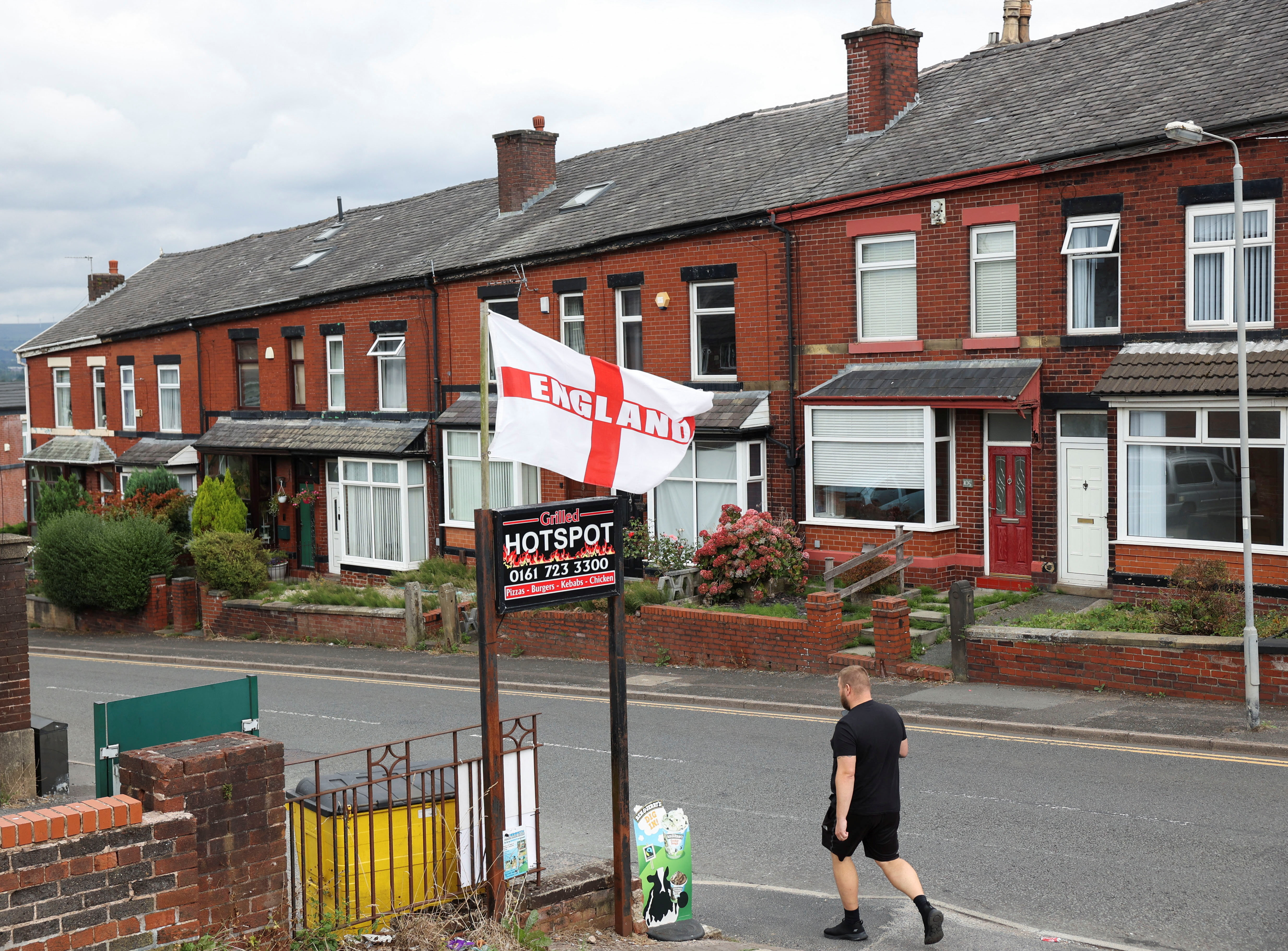 A man walks past a flag bearing the colours of England's St. George's Cross, flying above a residential street in Radcliffe, near Manchester,
