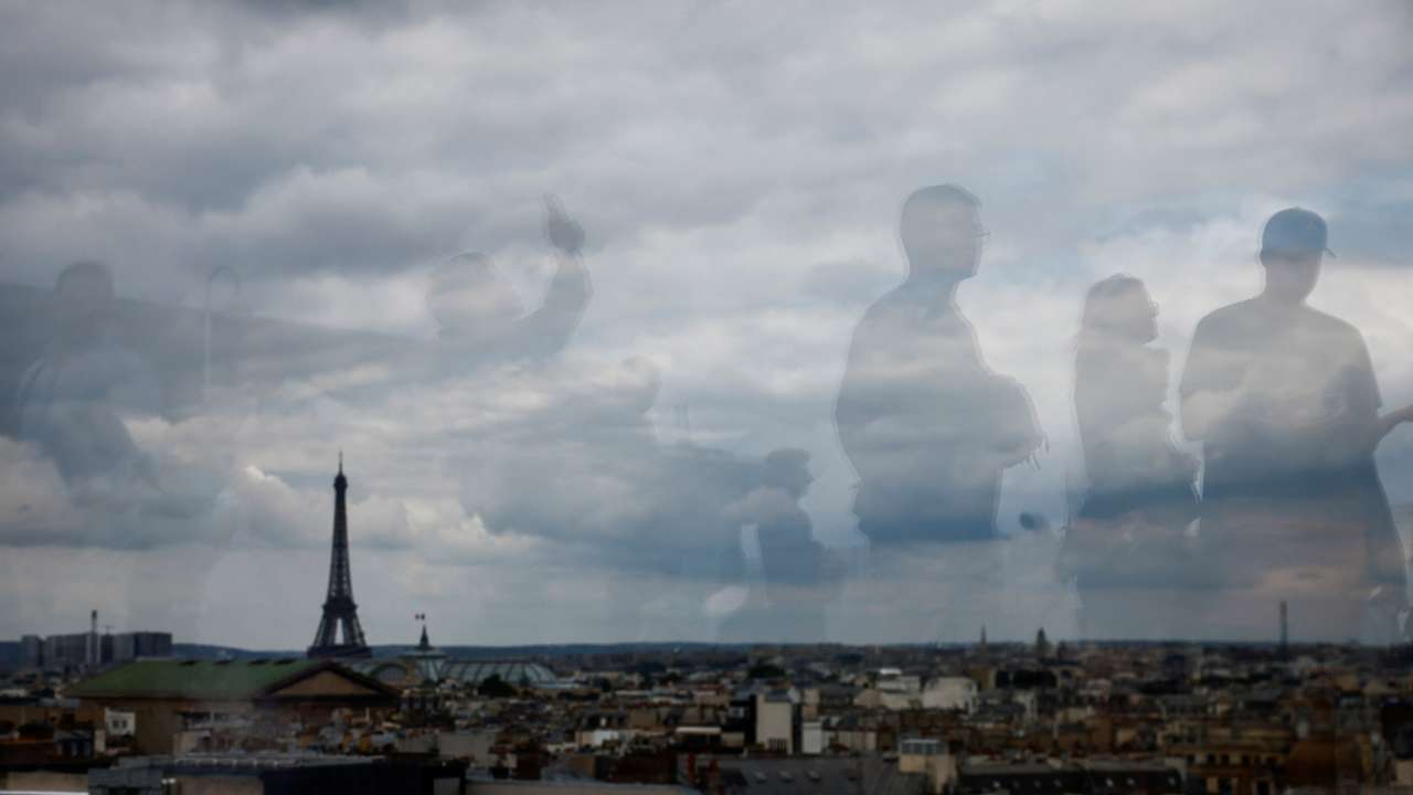 Tourists are reflected on the balustrade as they look at the Eiffel Tower and the Paris skyline from the rooftop of the Galeries Lafayette department store on a cloudy day in Paris