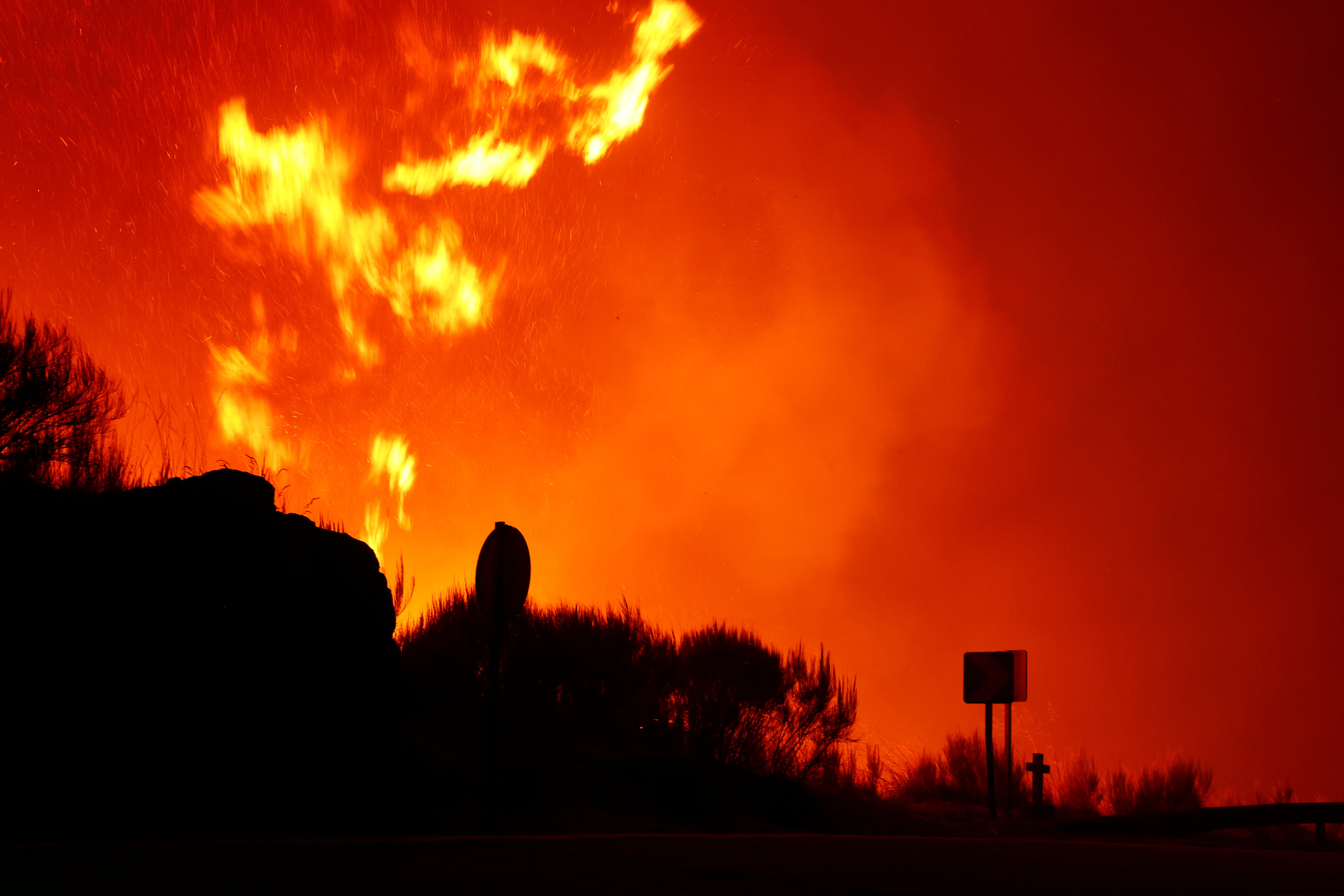 A cross on the road is silhouetted, as flames and smoke coming from a wildfire rise near Povoa de Montemuro