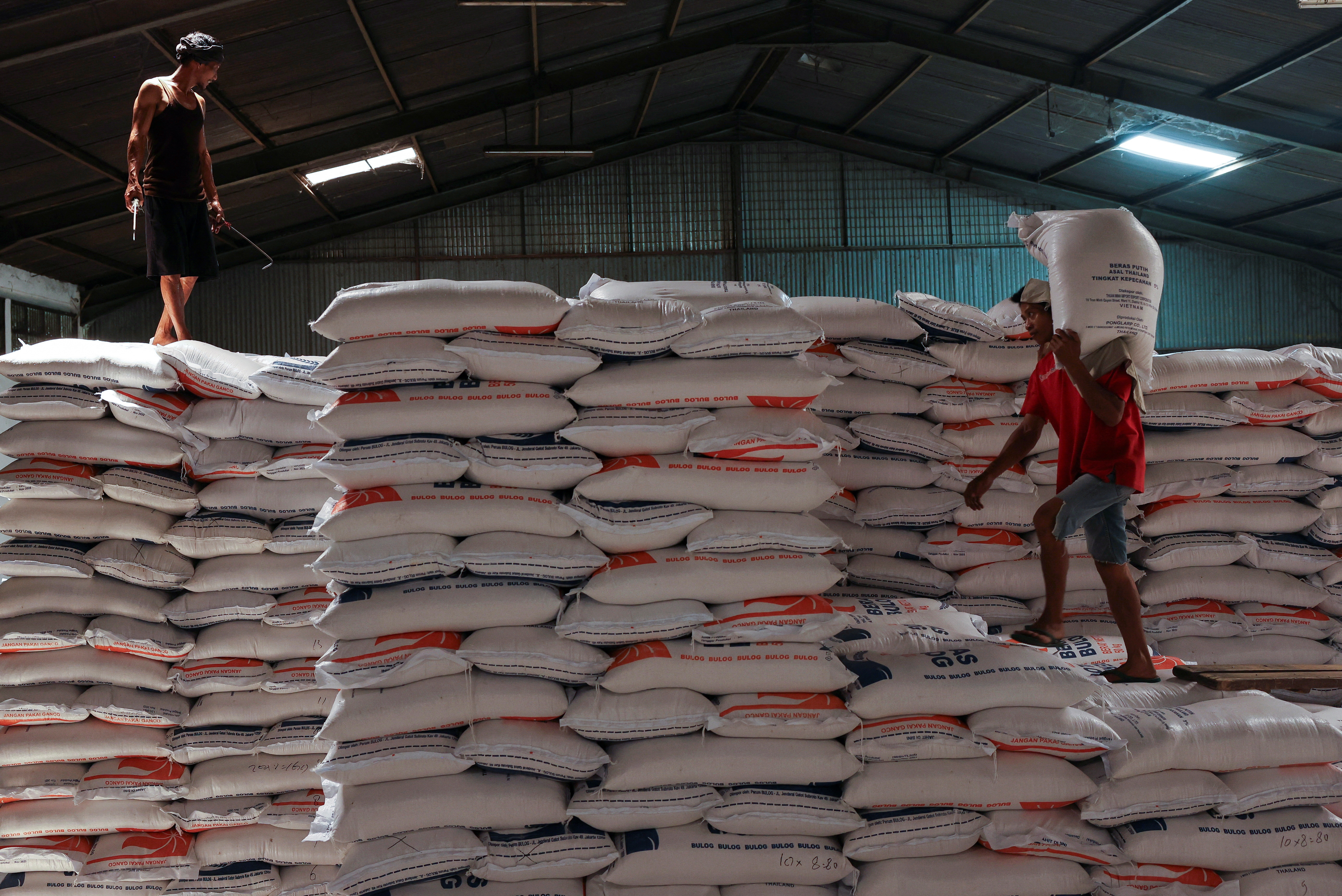 A worker stands on a pile of rice sacks as the other worker carries a sack of rice at the warehouse in Jakarta