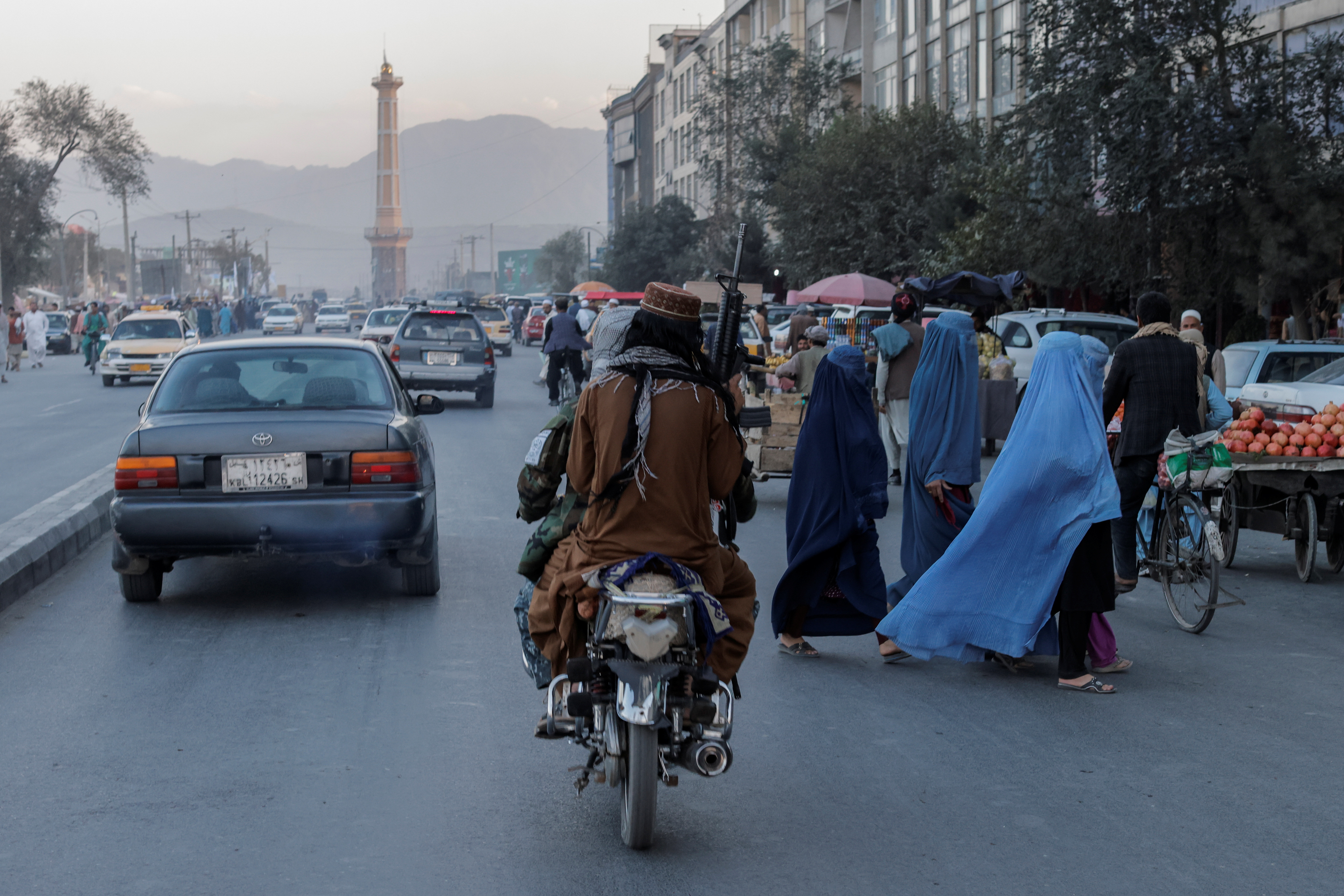 A group of women wearing burqas crosses the street as members of the Taliban drive past
