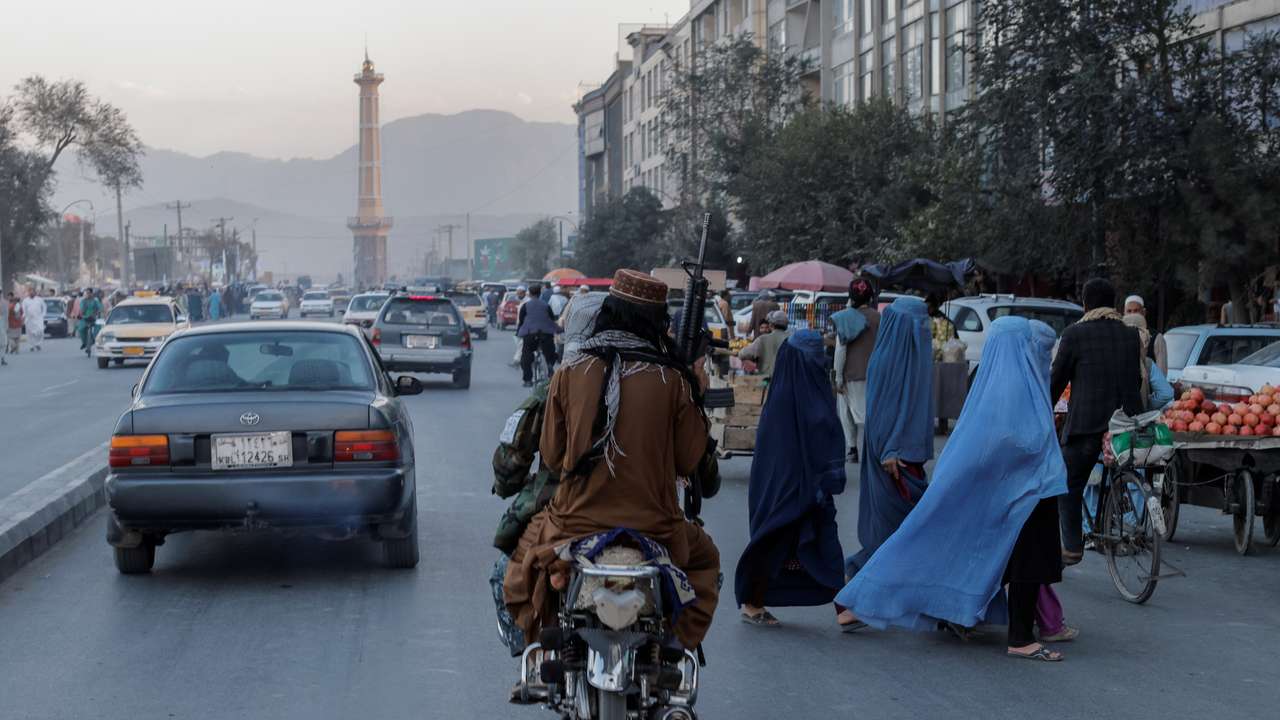 A group of women wearing burqas crosses the street as members of the Taliban drive past