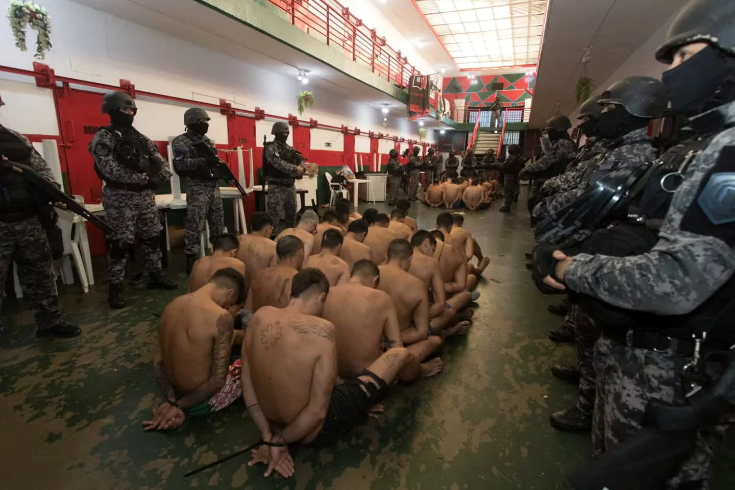 Half-naked inmates with their hands tied sit on the floor surrounded by armed police officers