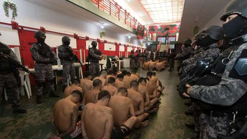 Half-naked inmates with their hands tied sit on the floor surrounded by armed police officers