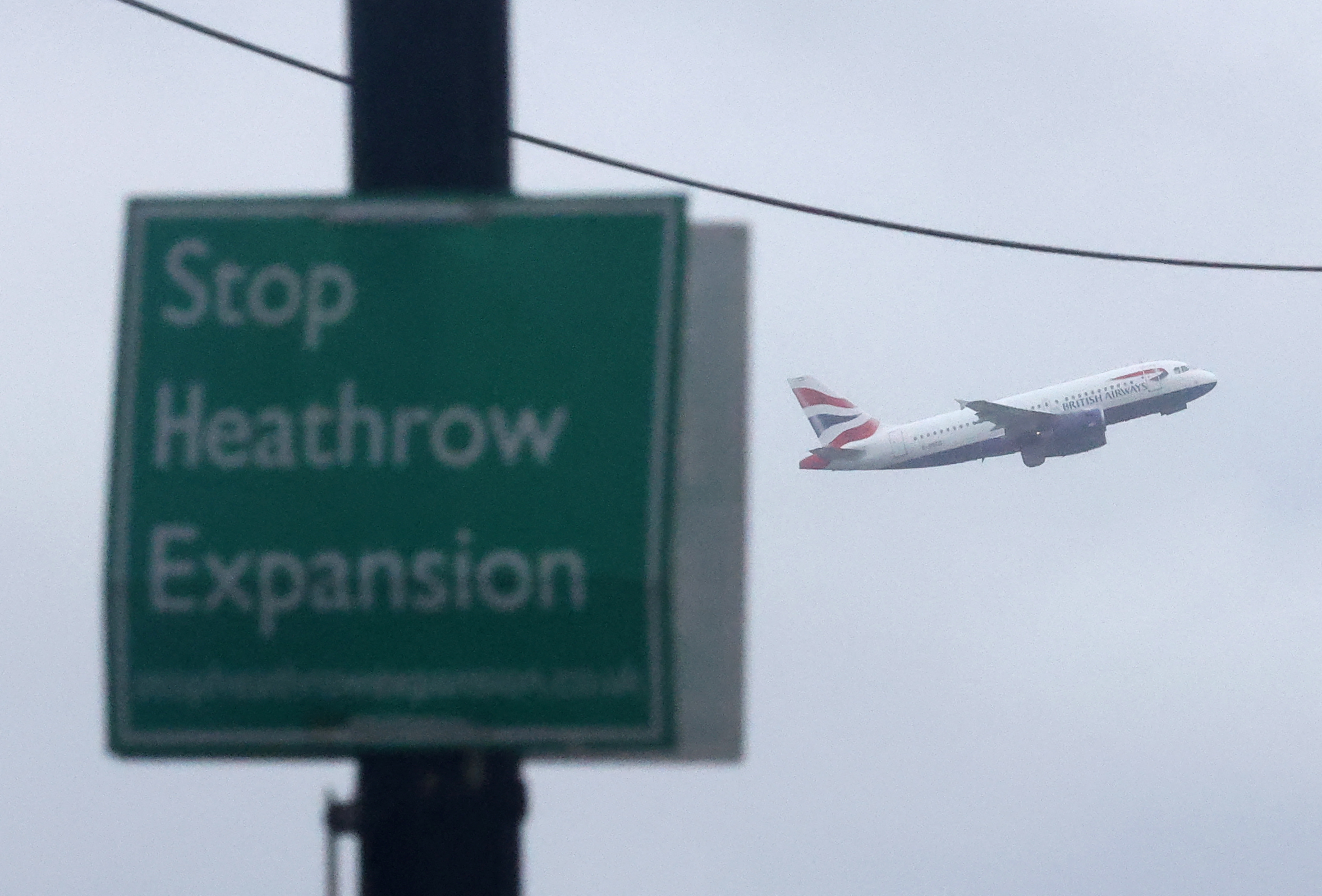Campaign sign opposing airport development seen attached to lamp post as British Airways passenger plane takes off from Heathrow Airport, Harmondsworth, west London