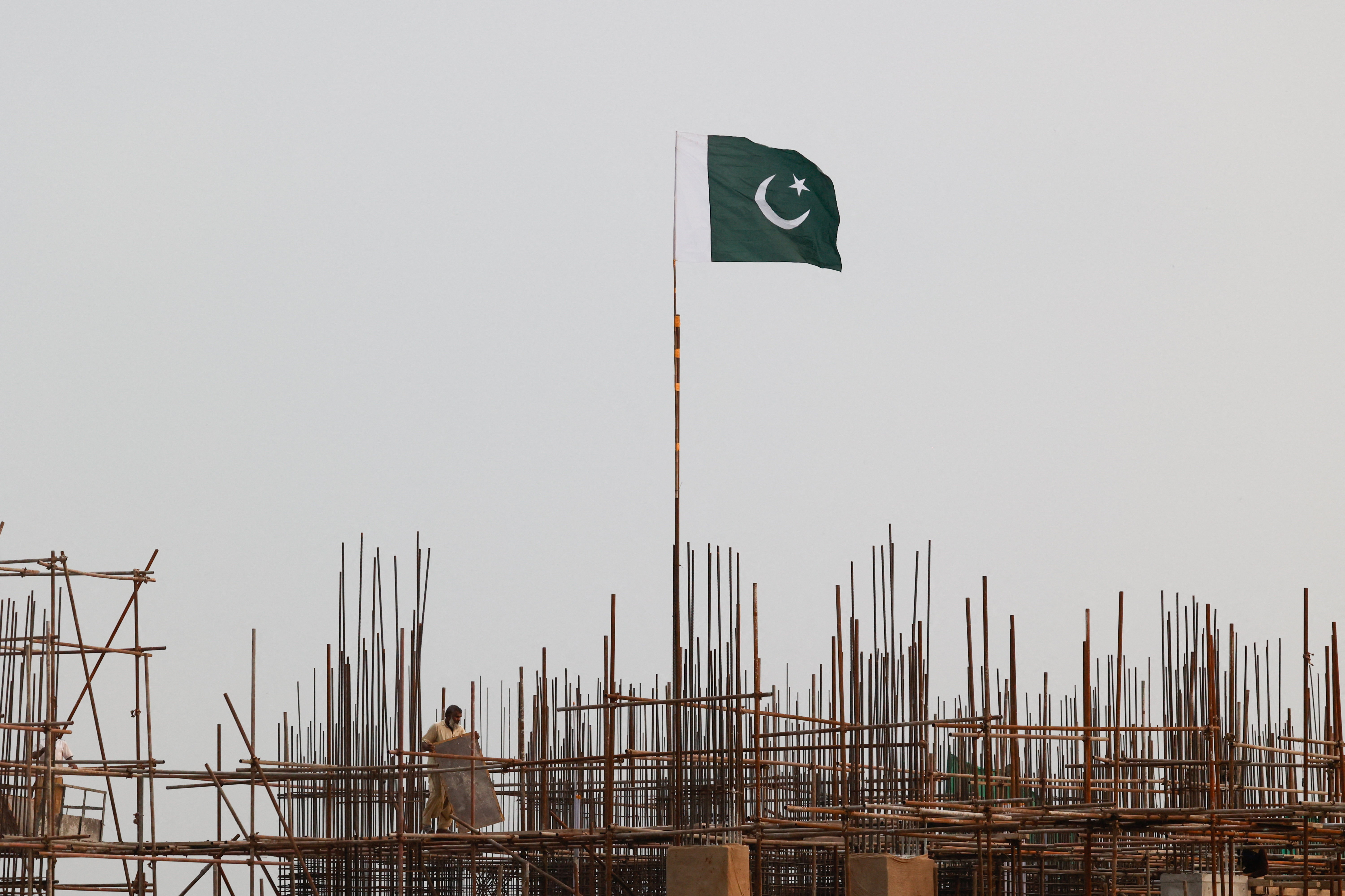 A Pakistani flag flutters over iron scaffoldings as a labourer works at a construction site on the eve of Pakistan's Independence Day celebrations, in Lahore