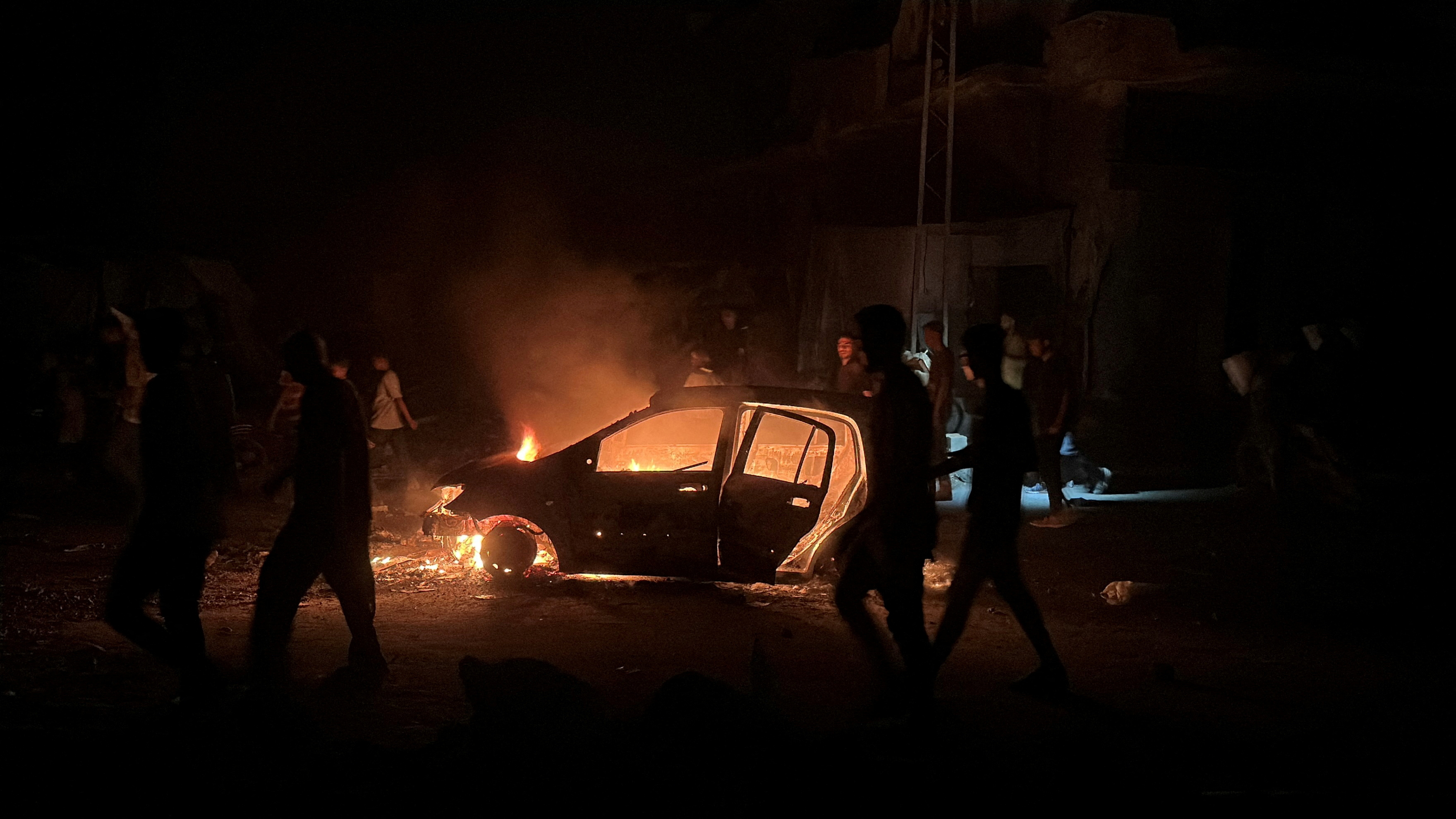 Palestinians walk past a burning car hit in an Israeli strike, in Khan Younis, in the southern Gaza Strip