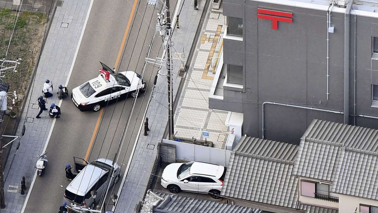 Police officers take cover behind cars outside the post office where a suspected gunman has taken people hostage after injuring two at a hospital, in Warabi,