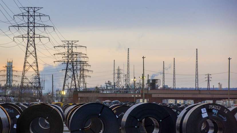 Coils of rolled steel sit in an industrial yard with transmission towers and smokestacks in the background at dusk in Hamilton