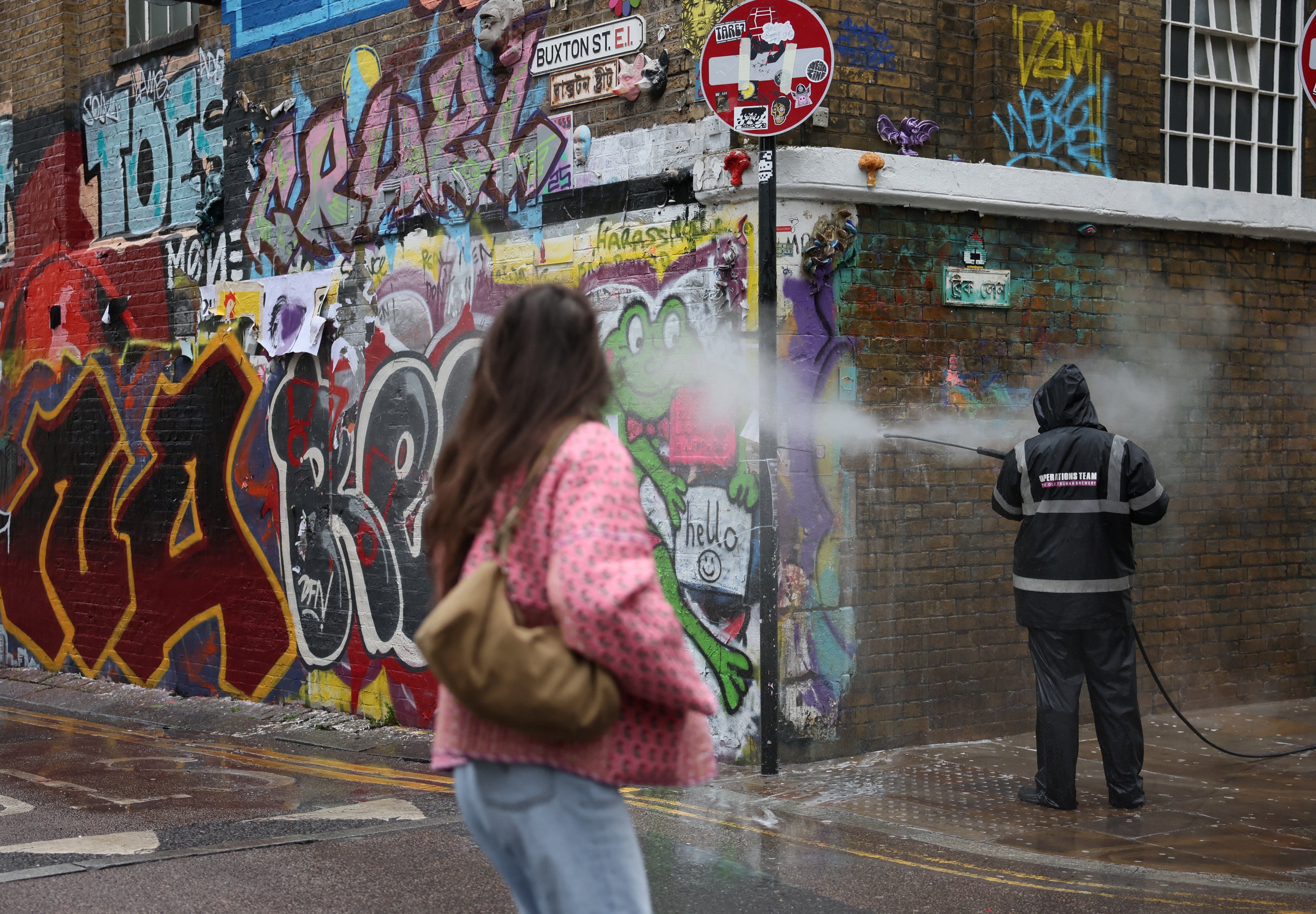 A woman walks past a worker cleaning graffiti from a wall in Brick Lane, east London