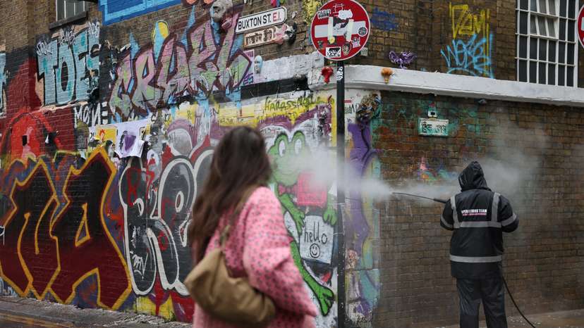 A woman walks past a worker cleaning graffiti from a wall in Brick Lane, east London