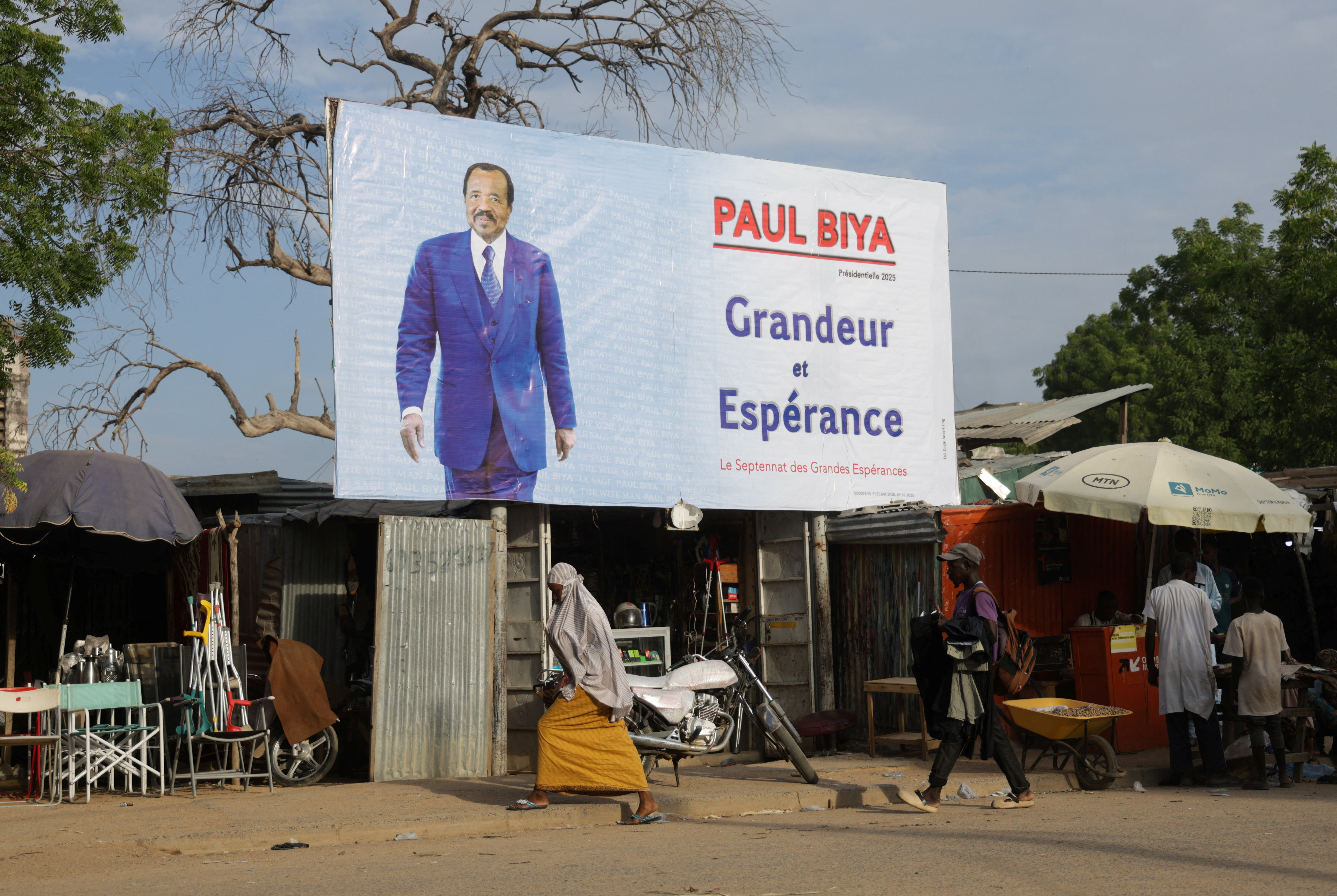 People walk past an election campaign poster for the incumbent President Paul Biya in Maroua, Cameroon