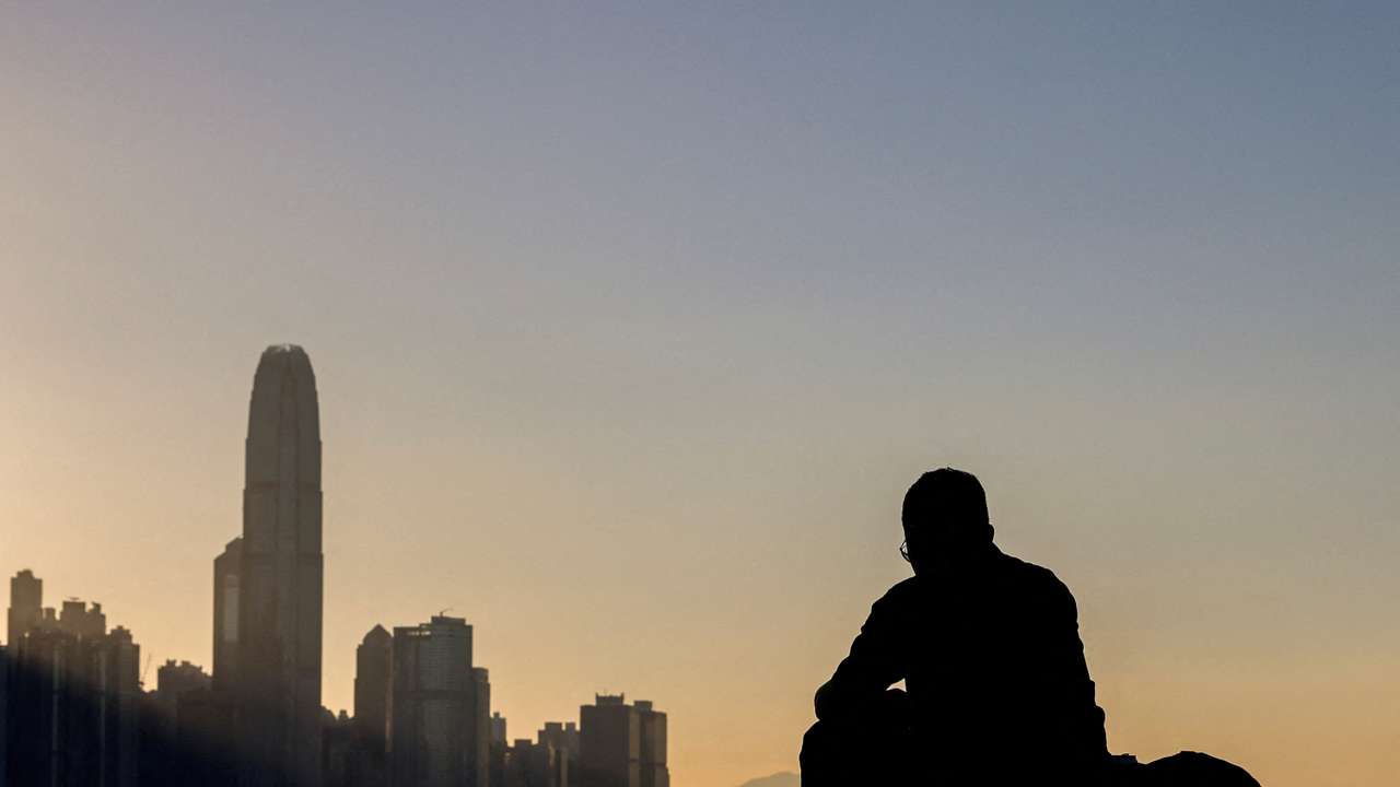 A man sits by the waterfront with the skyline buildings across Victoria Harbor in Hong Kong