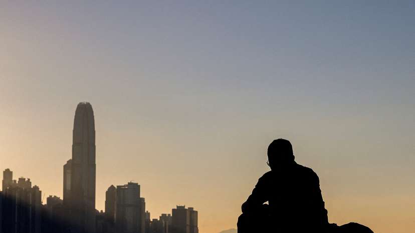 A man sits by the waterfront with the skyline buildings across Victoria Harbor in Hong Kong