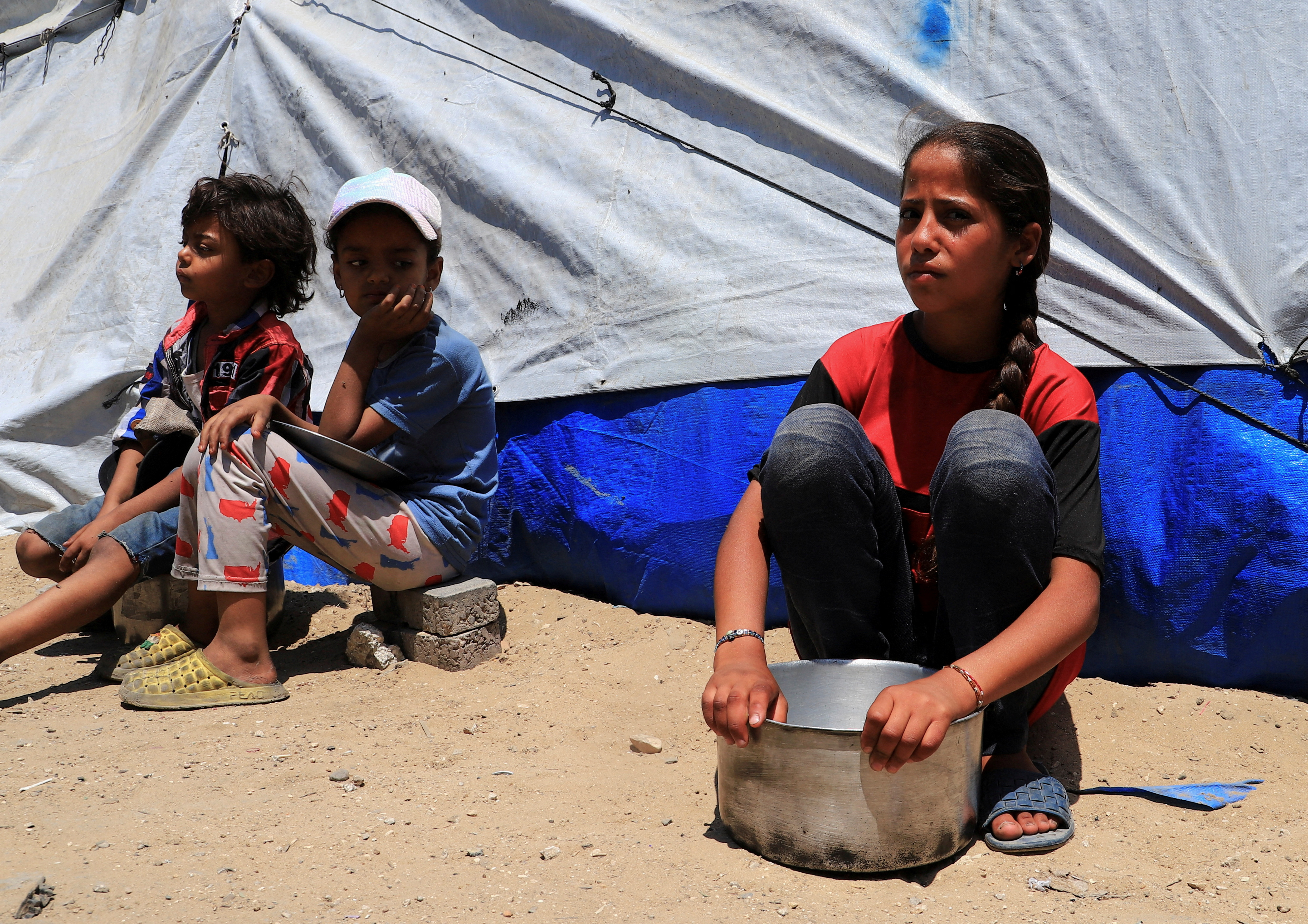 FILE PHOTO: Displaced Palestinian children, one with an empty pot, sit while waiting to receive food from a charity kitchen, in Gaza City