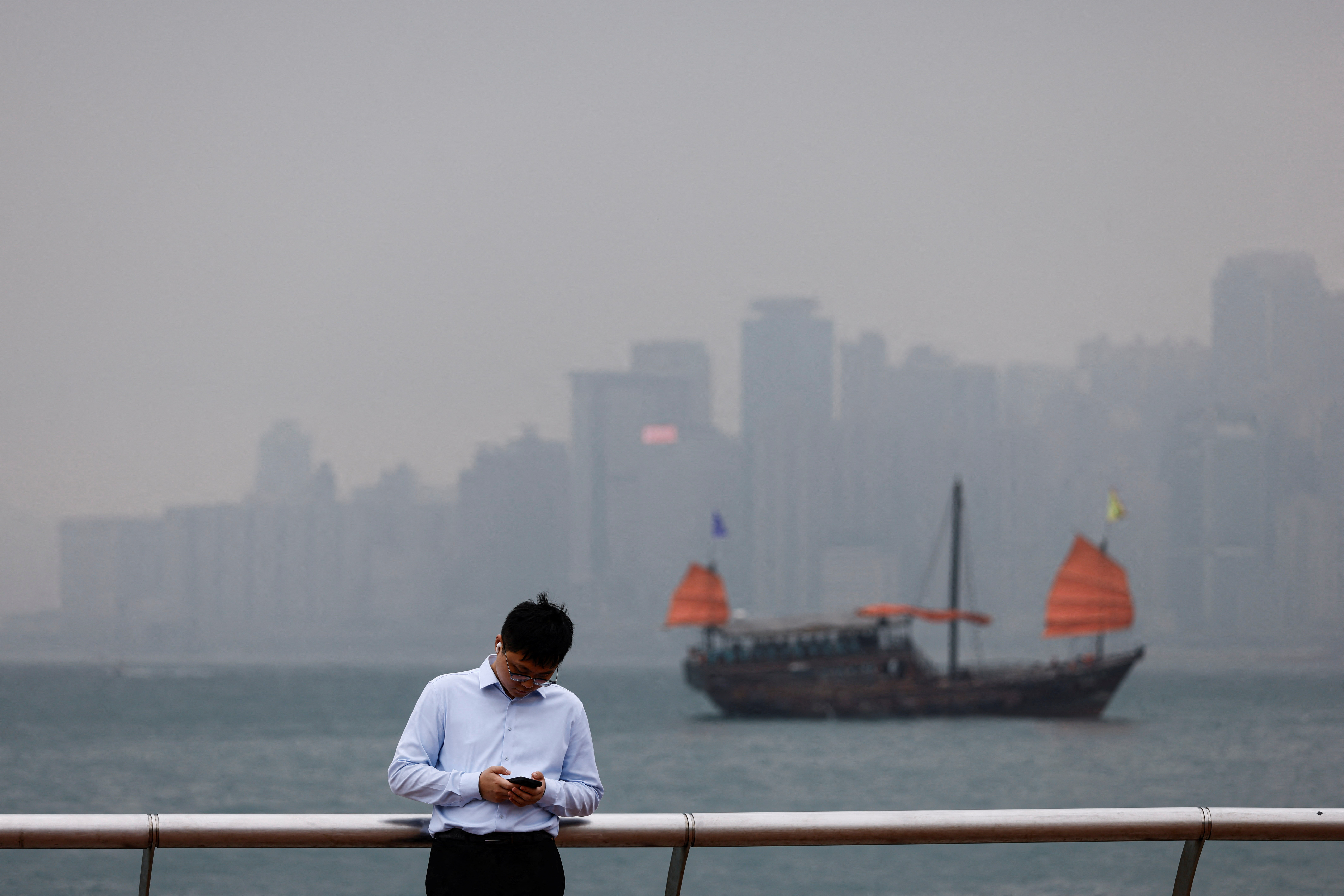 A man checks his phone at the waterfront, with a tourist junk boat in the backdrop, on a foggy day in Hong Kong
