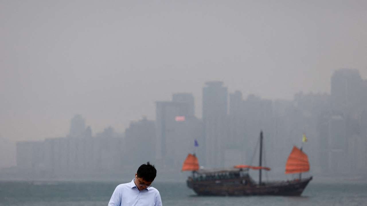 A man checks his phone at the waterfront, with a tourist junk boat in the backdrop, on a foggy day in Hong Kong
