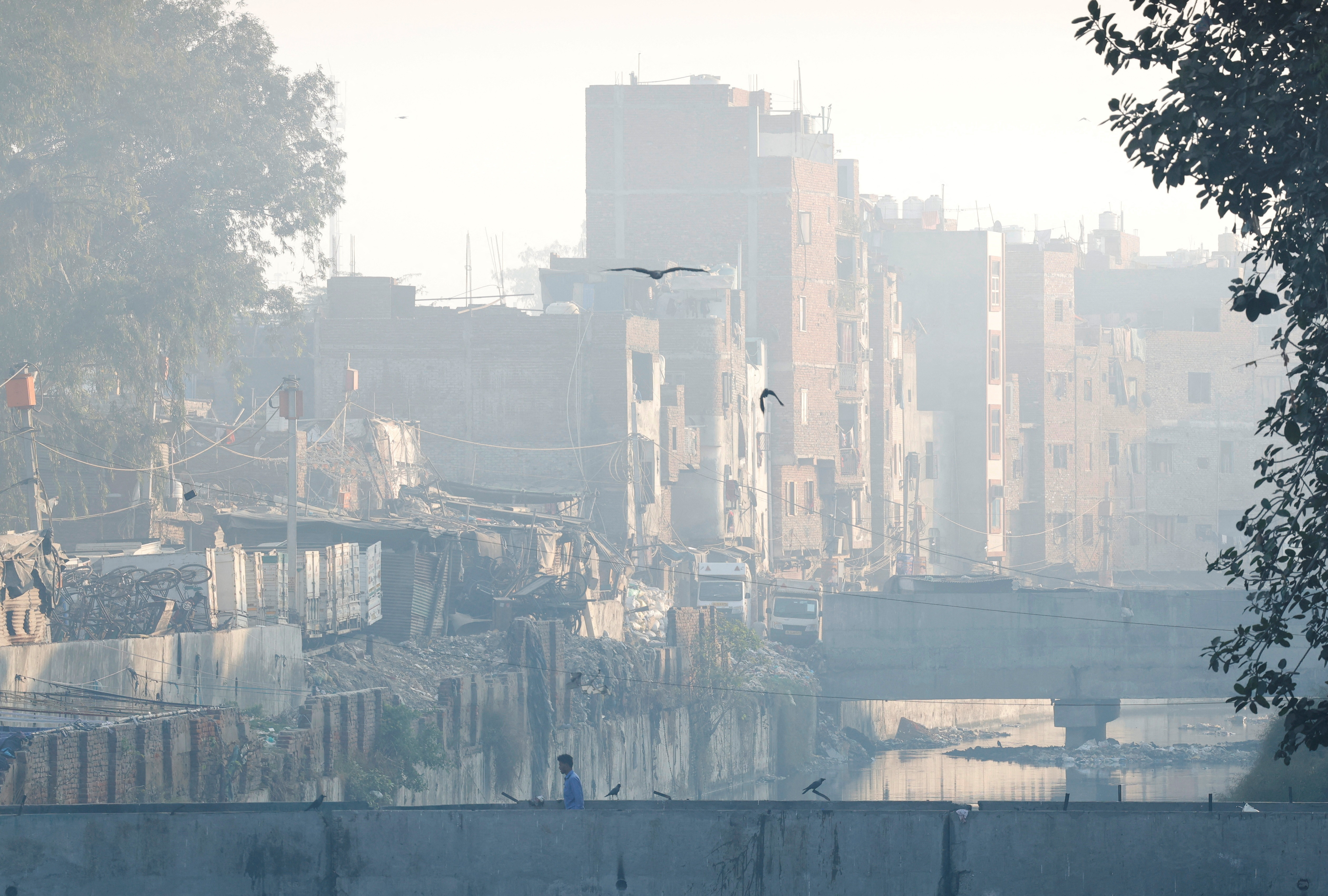 A man walks on a footbridge across an open drainage channel as the sky is enveloped in smog, in New Delhi