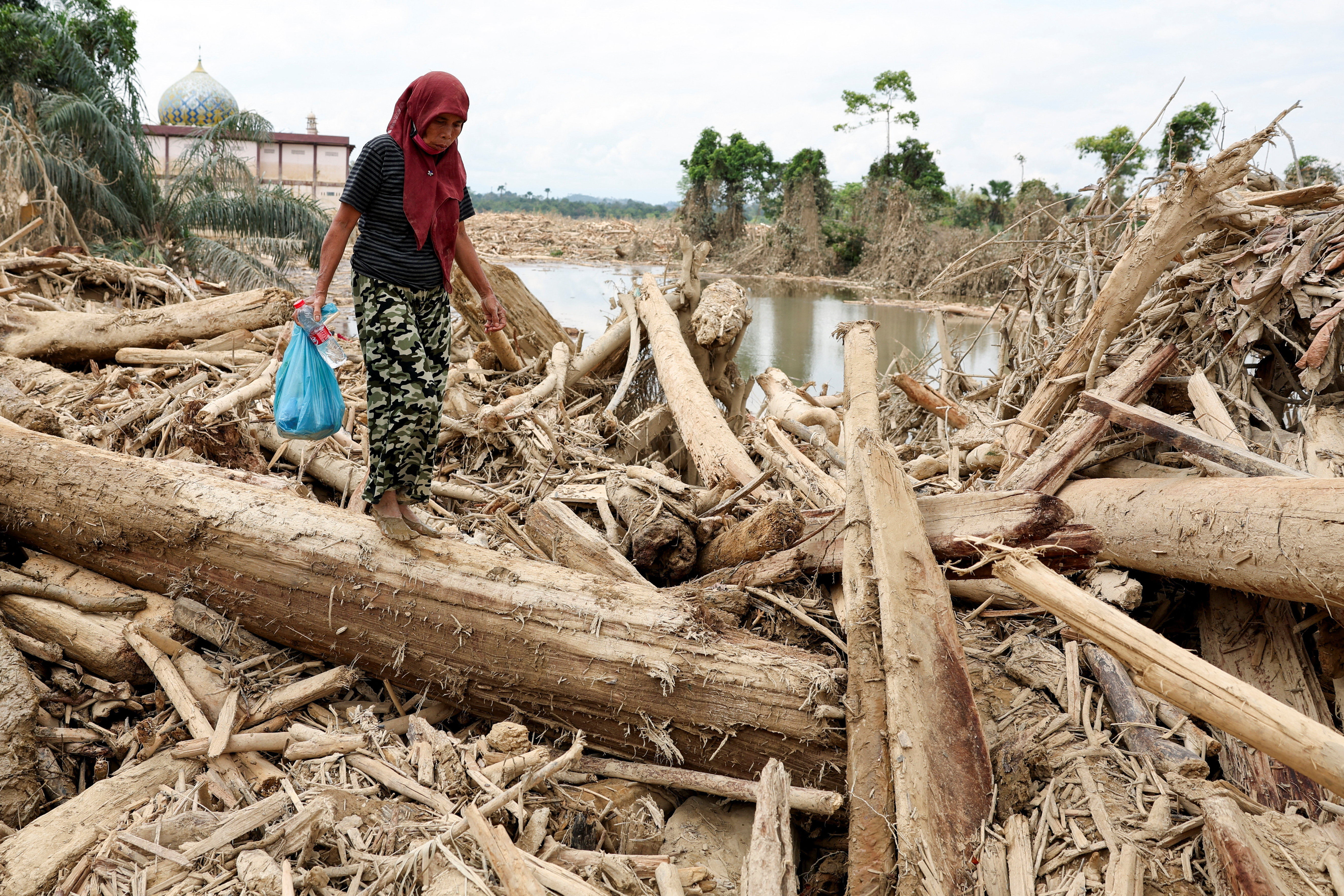 A survivor carries relief supplies while walking through tree trunks that were stranded in an area affected by a deadly flash flood following heavy rains in Karang Baru