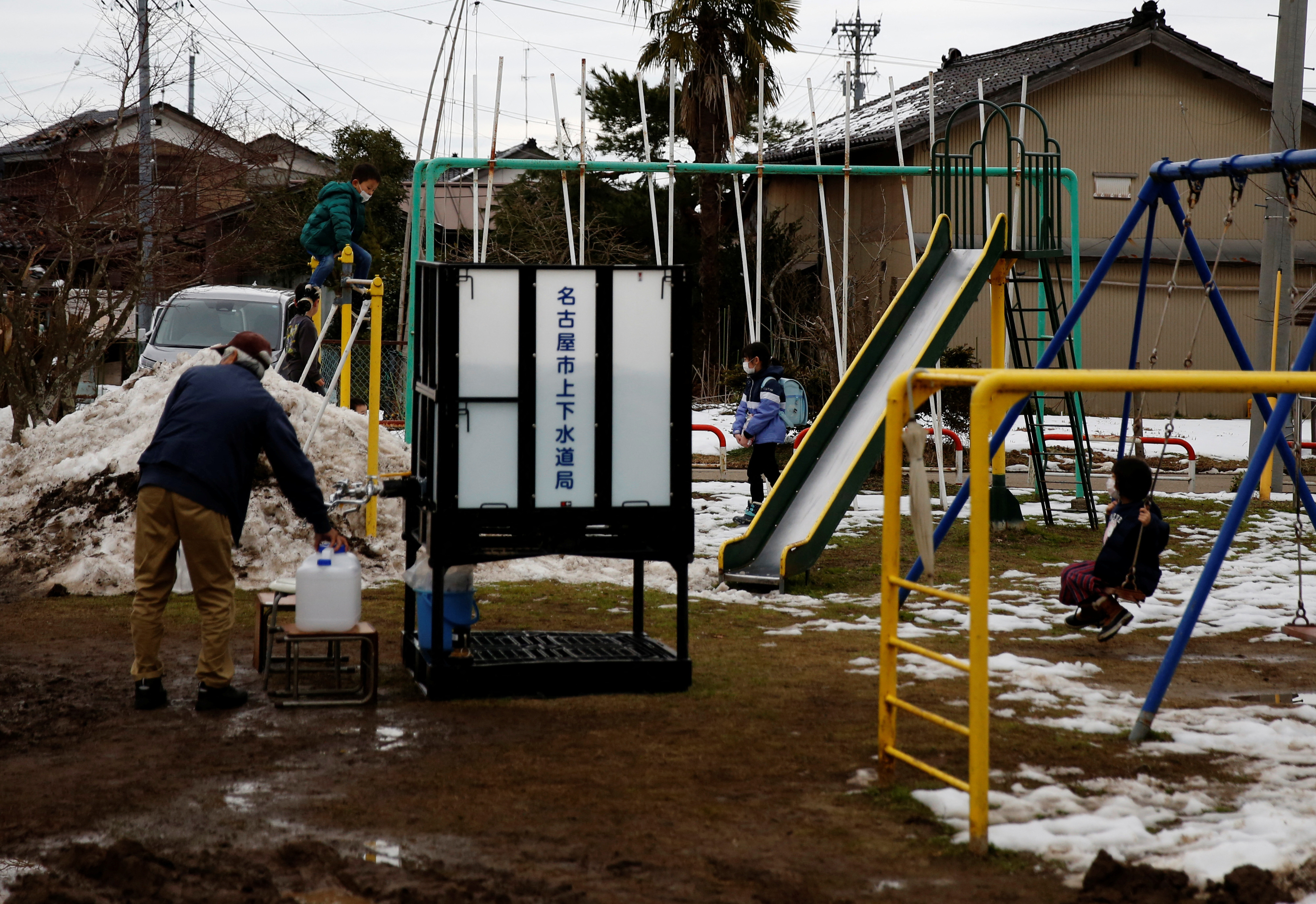 Children play in the playground as a local resident refills a bottle from the communal water tank for Noto earthquake survivors in Suzu