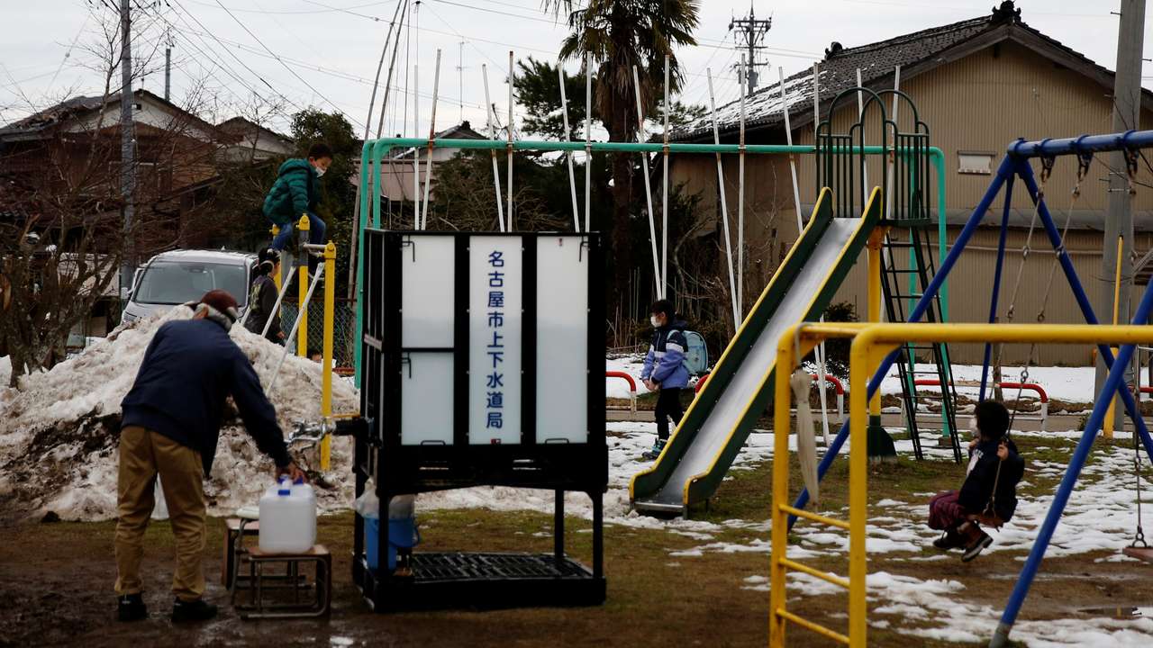 Children play in the playground as a local resident refills a bottle from the communal water tank for Noto earthquake survivors in Suzu