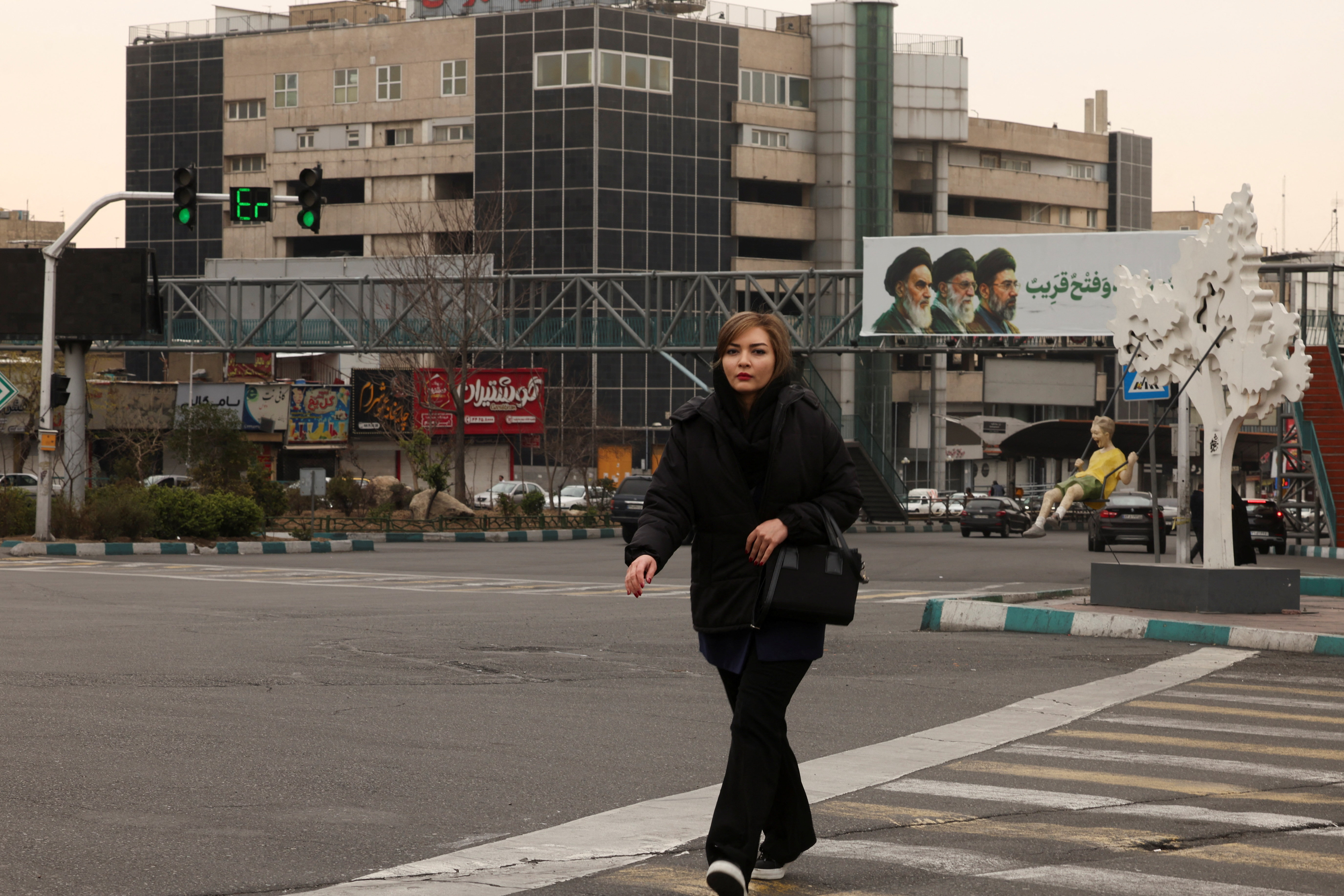 A woman walks across the street, with a banner of all three leaders of Iran, late Ruhollah Khomeini, late Ayatollah Ali Khamenei, and Mojtaba Khamenei in the background, in Tehran