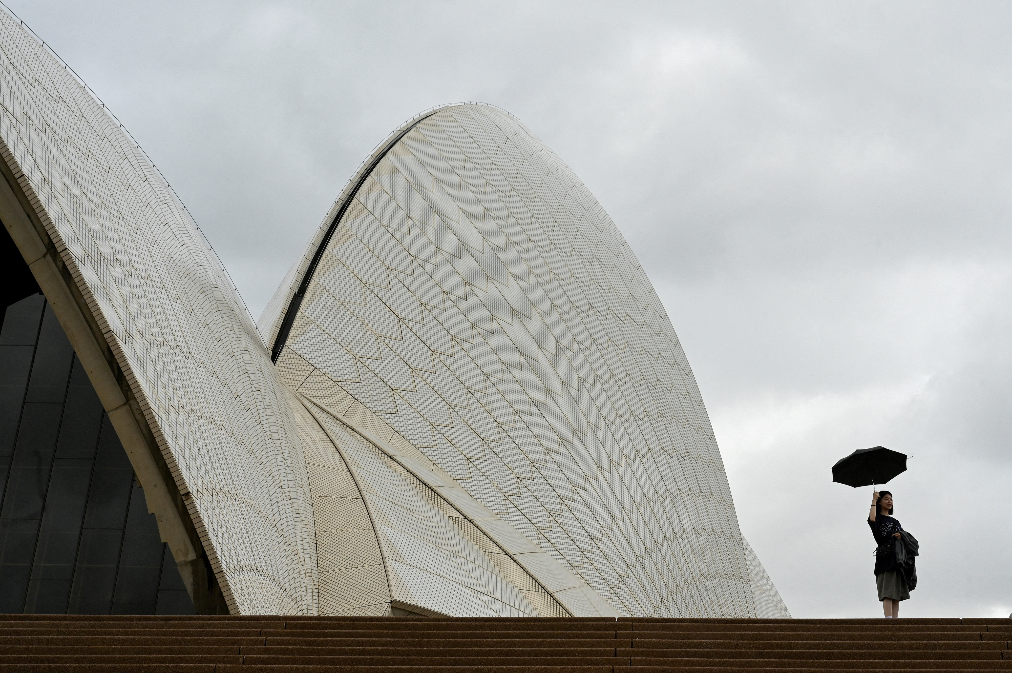 A woman holds up an umbrella at the Sydney Opera House during cloudy and rainy weather conditions in Sydney