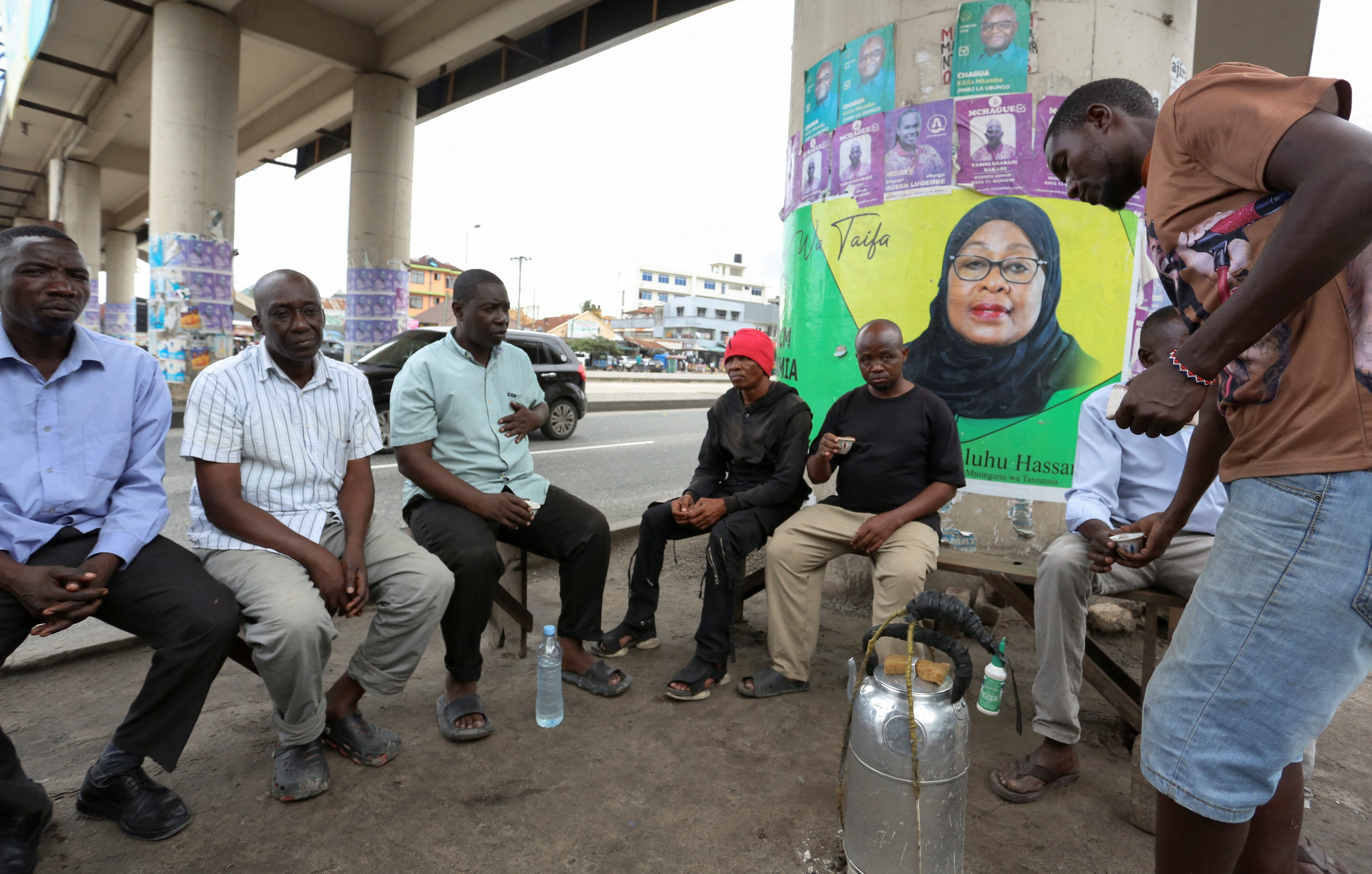 People sit under a foot bridge with an electoral campaign poster of Tanzania's President Samia Suluhu Hassan of the ruling CCM ahead of the general elections in Ilala district of Dar es Salaam