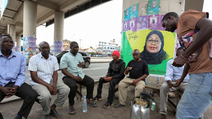 People sit under a foot bridge with an electoral campaign poster of Tanzania's President Samia Suluhu Hassan of the ruling CCM ahead of the general elections in Ilala district of Dar es Salaam