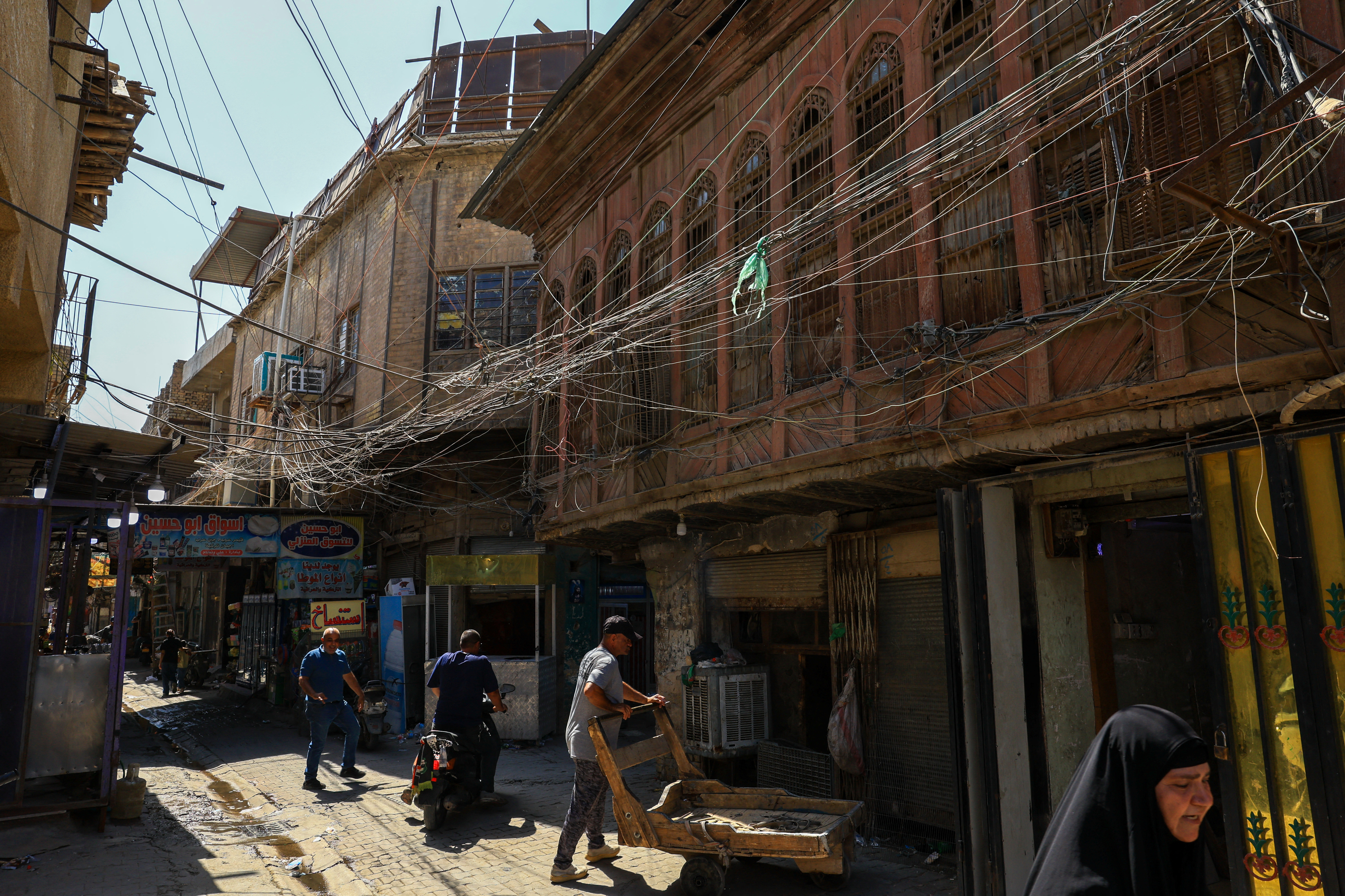 People walk along an old street as a tangled web of electrical wires from a generator supplying homes with electricity hangs above buildings, in Baghdad