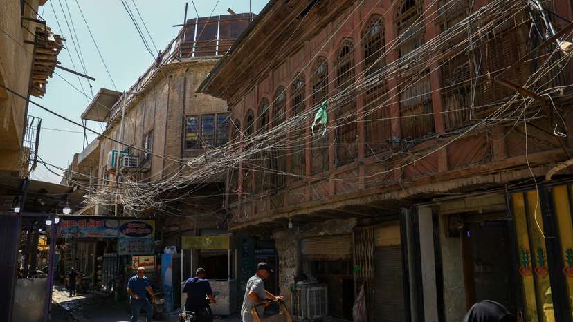 People walk along an old street as a tangled web of electrical wires from a generator supplying homes with electricity hangs above buildings, in Baghdad