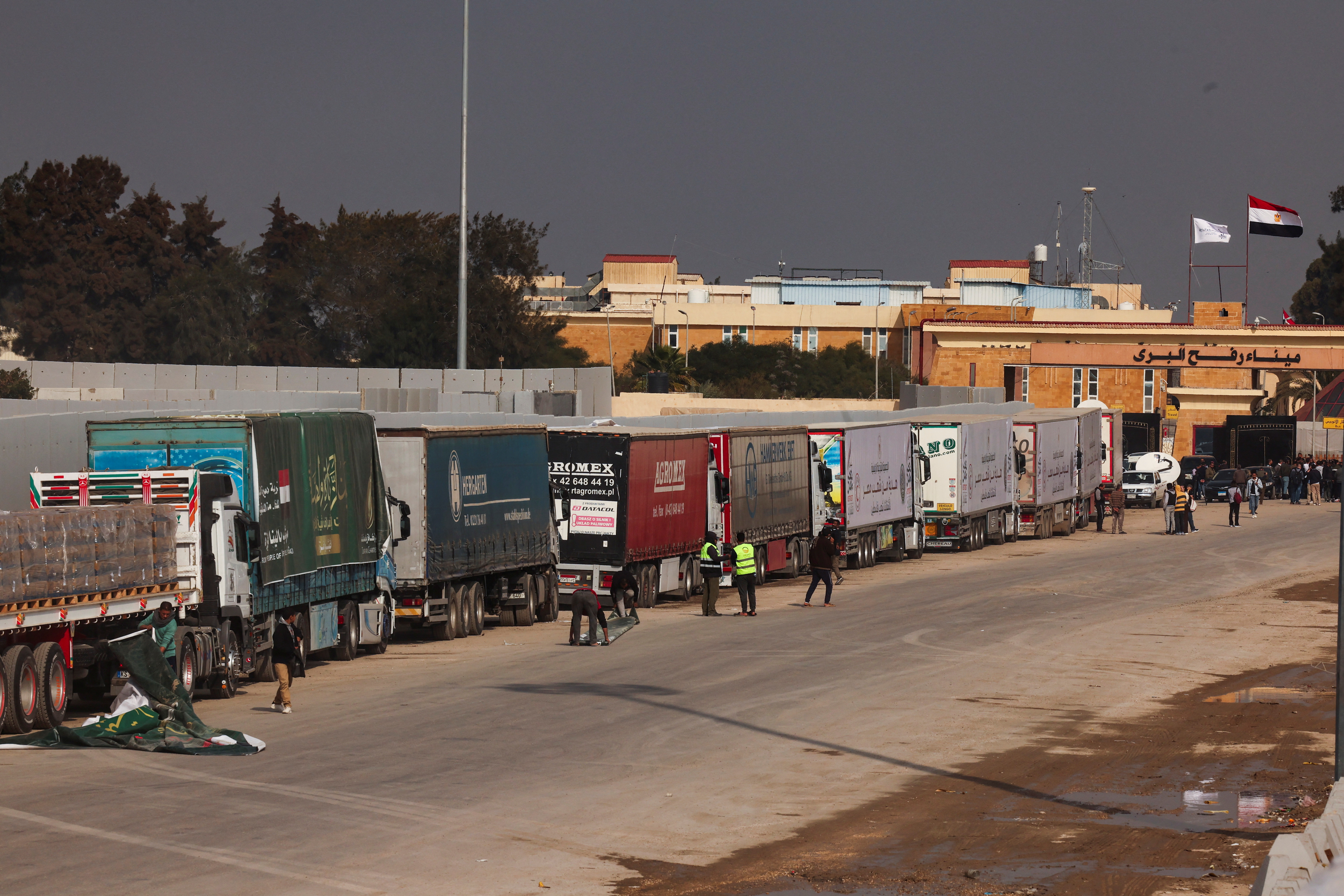 People stand next to trucks carrying humanitarian aid, at the Rafah border crossing to enter Gaza
