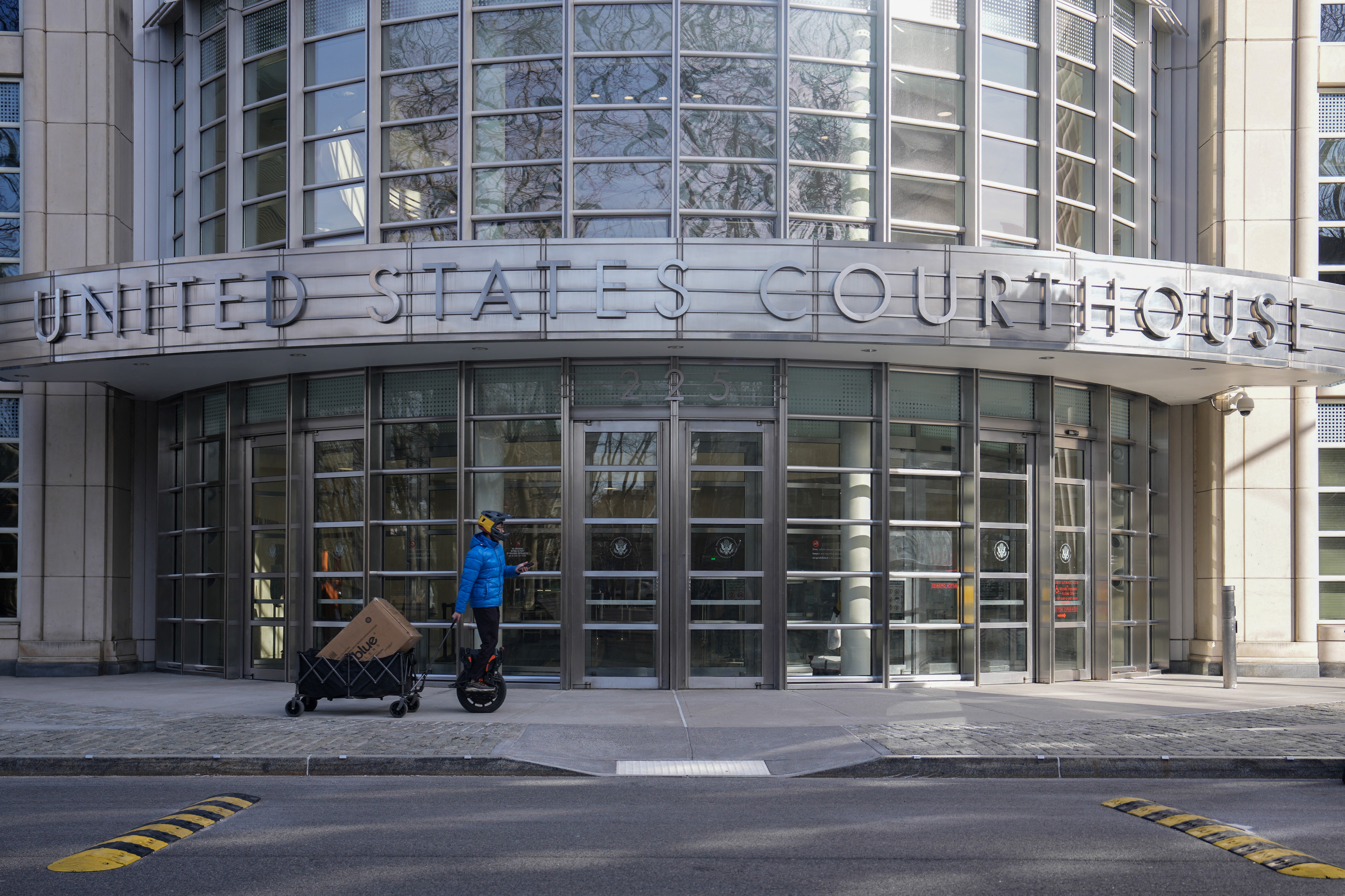 A person rides an electric unicycle while pulling a wagon loaded with packages, in front of the Eastern District of New York Court Federal Court House in New York City