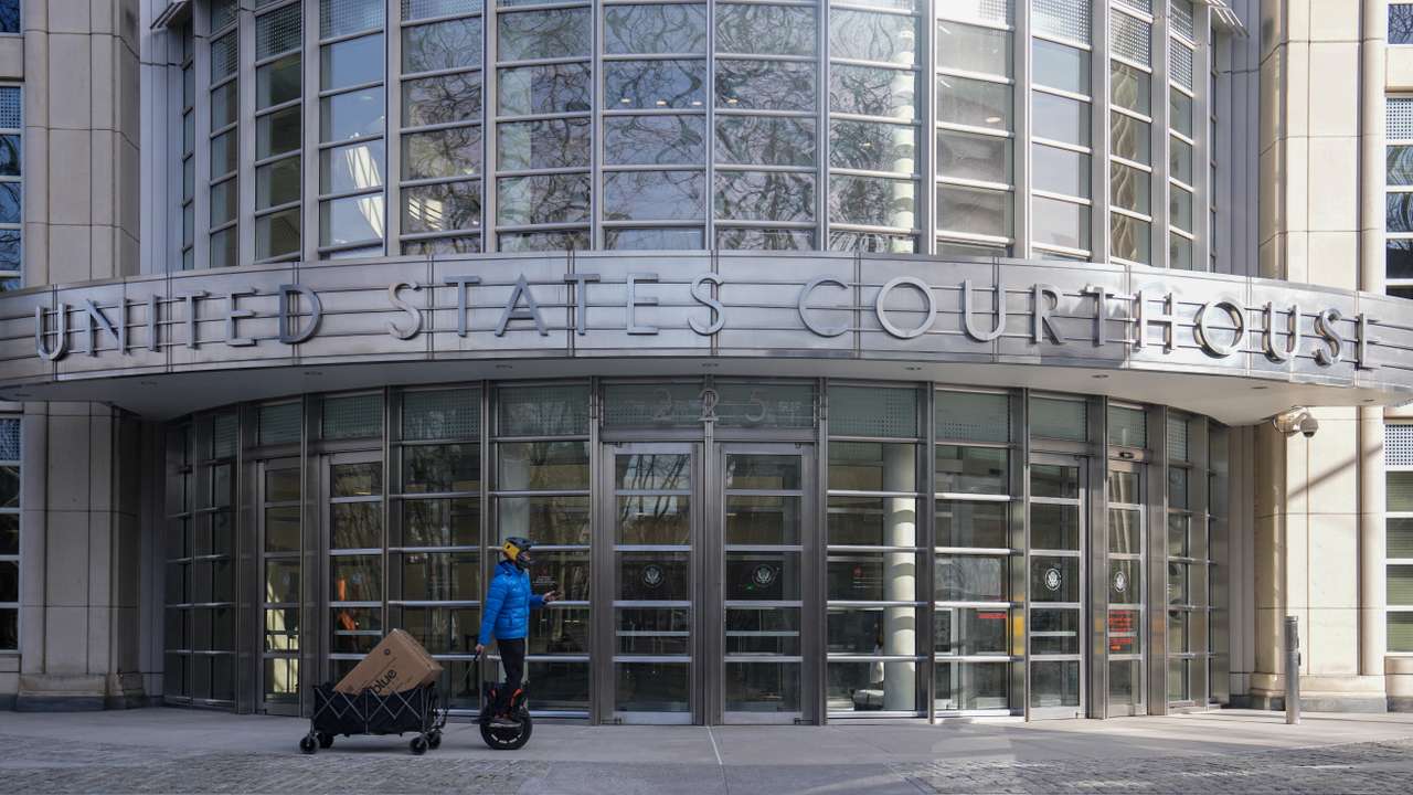 A person rides an electric unicycle while pulling a wagon loaded with packages, in front of the Eastern District of New York Court Federal Court House in New York City