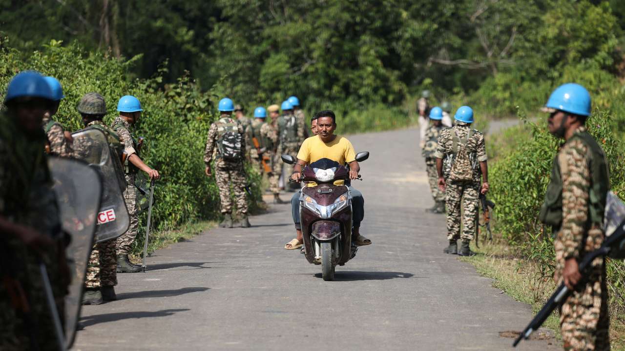 People ride a scooter past Central Reserve Police Force personnel standing guard before the funeral of Meiteis who were killed after ethnic violence broke out in Borobekra, Jiribam