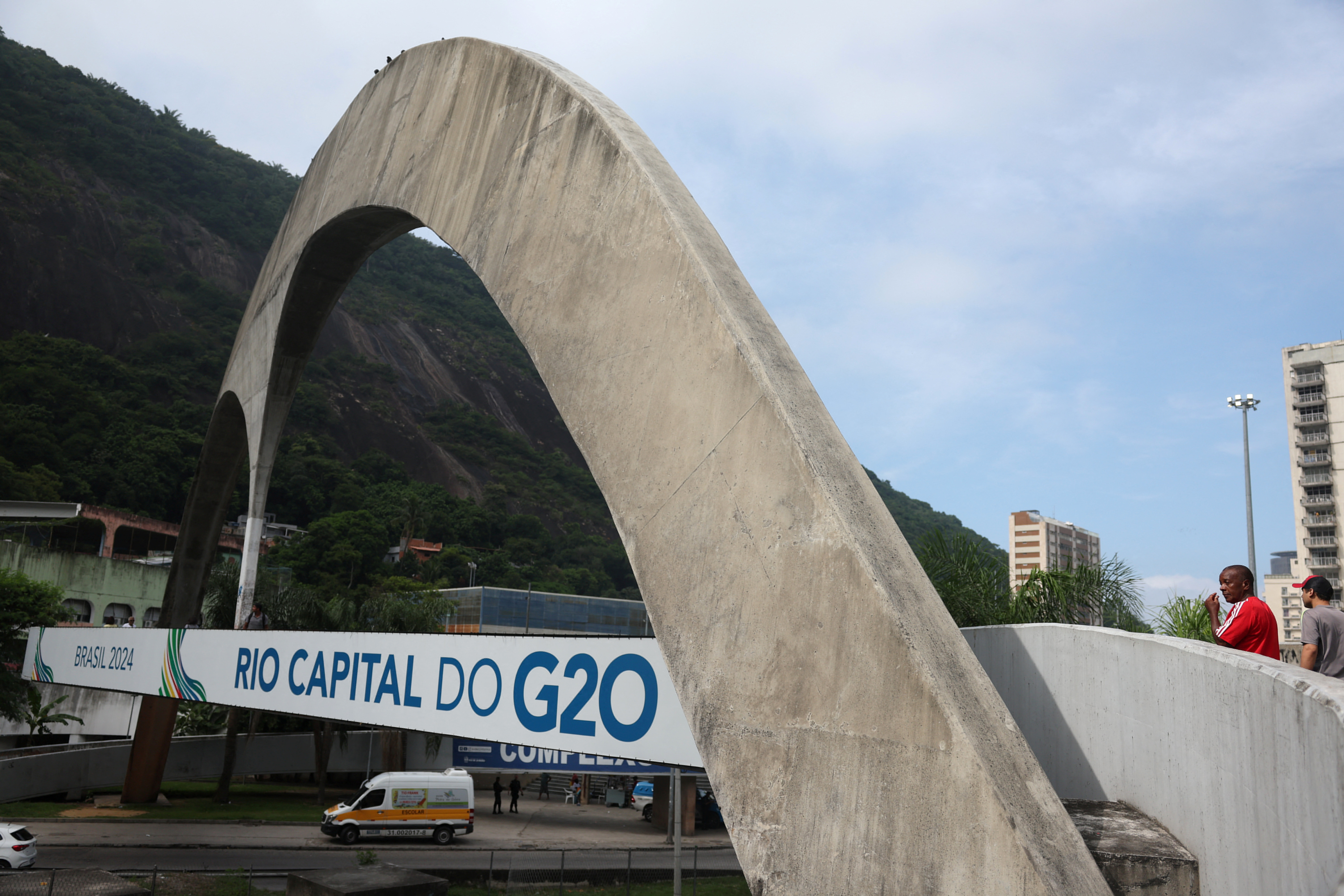 People walk next to a banner ahead of a meeting of foreign ministers, as part of Brazil's presidency of the G20 group  of advanced economies, in the Rocinha slum in Rio de Janeiro