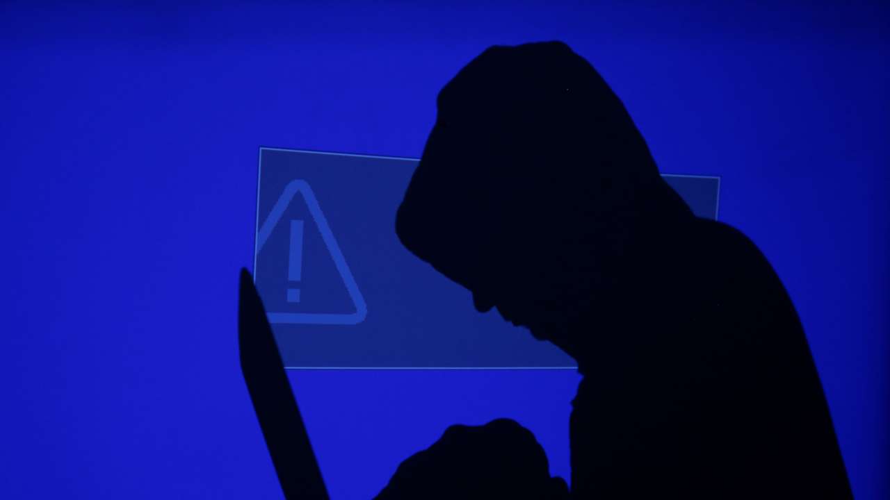 Hooded man holds laptop computer as blue screen with an exclamation mark is projected on him in this illustration picture