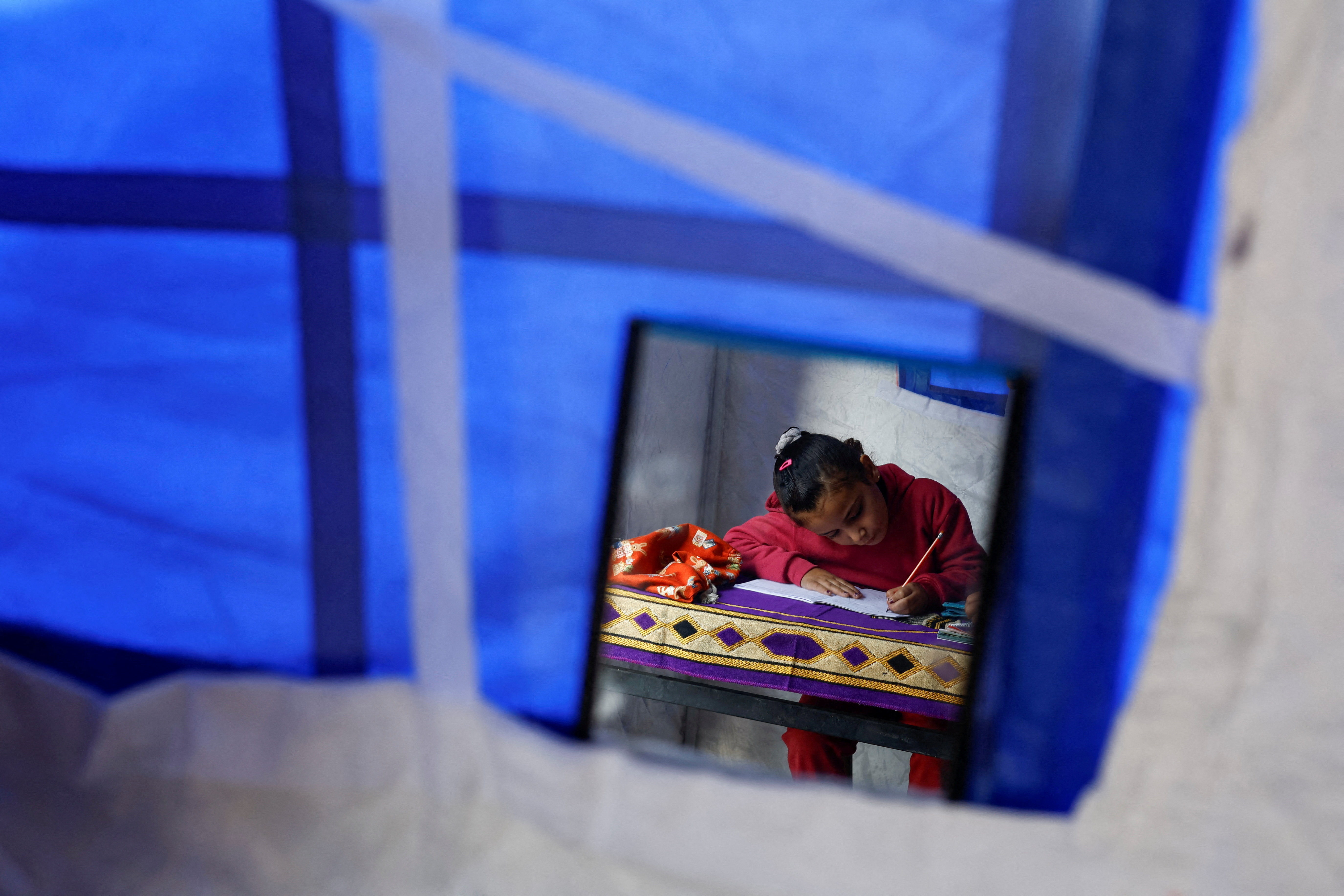 Displaced Palestinian girl Toulin Al-Hindi, is reflected in a hanging mirror as she does her homework inside her family's tent near the Israeli-designated "yellow line" in Beit Lahiya