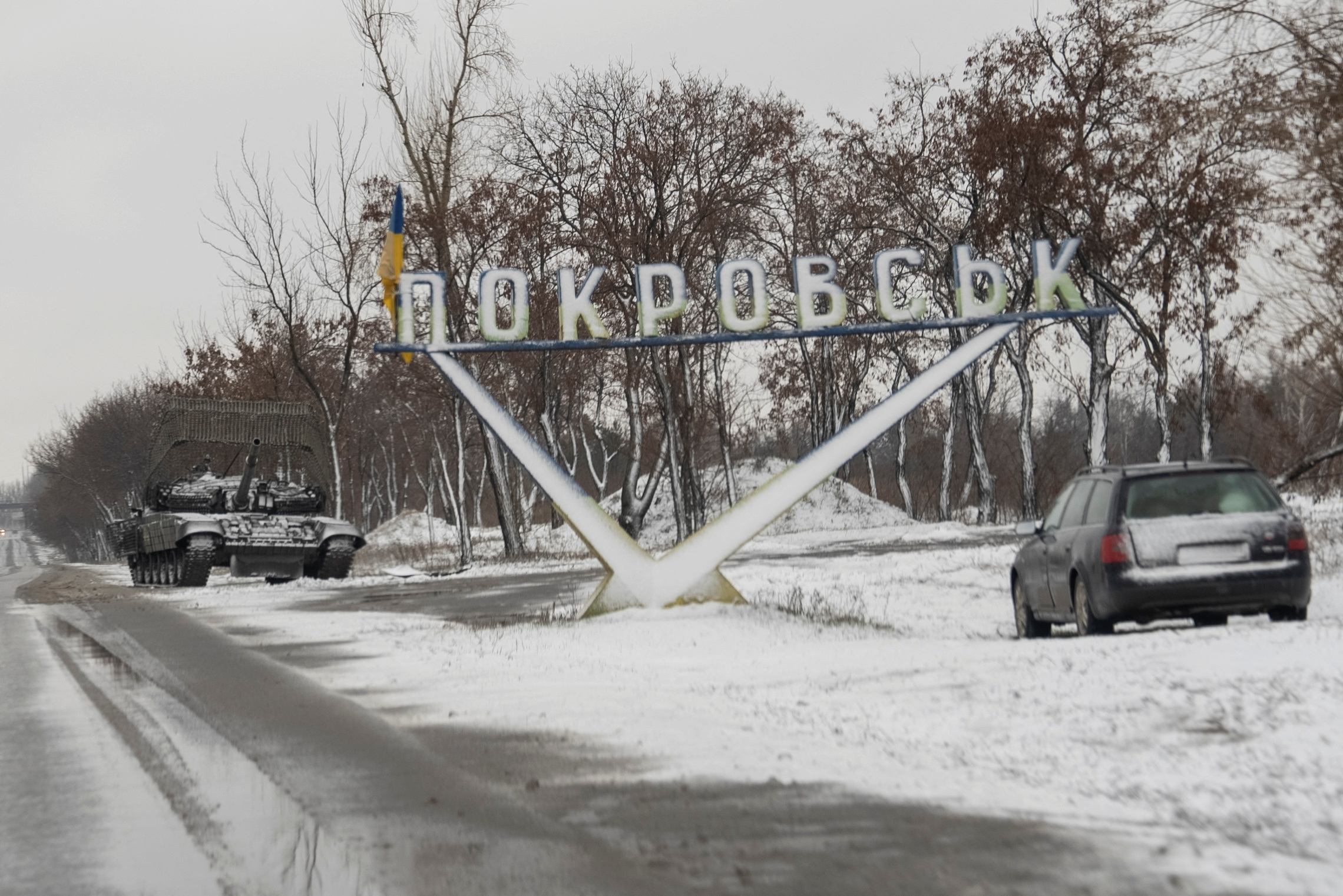 A Ukrainian tank and a car are parked near a road sign that reads, 'Pokrovsk’ in the town of Pokrovsk
