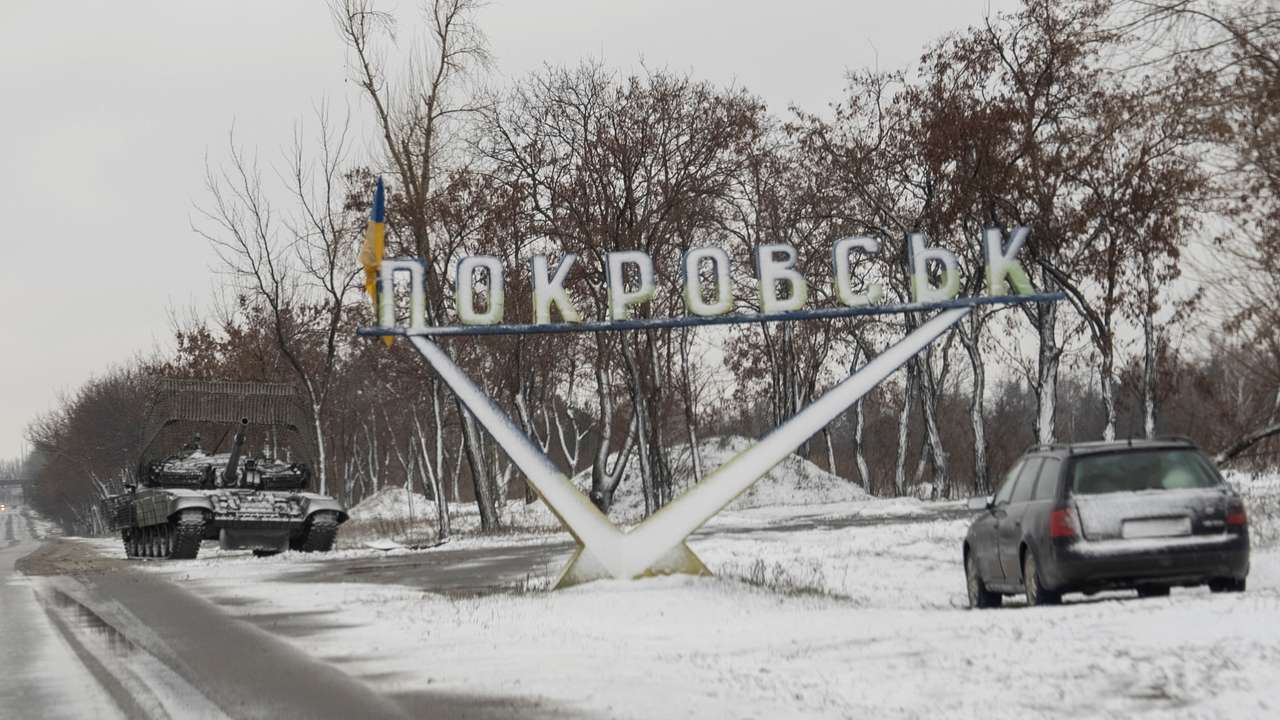 A Ukrainian tank and a car are parked near a road sign that reads, 'Pokrovsk’ in the town of Pokrovsk