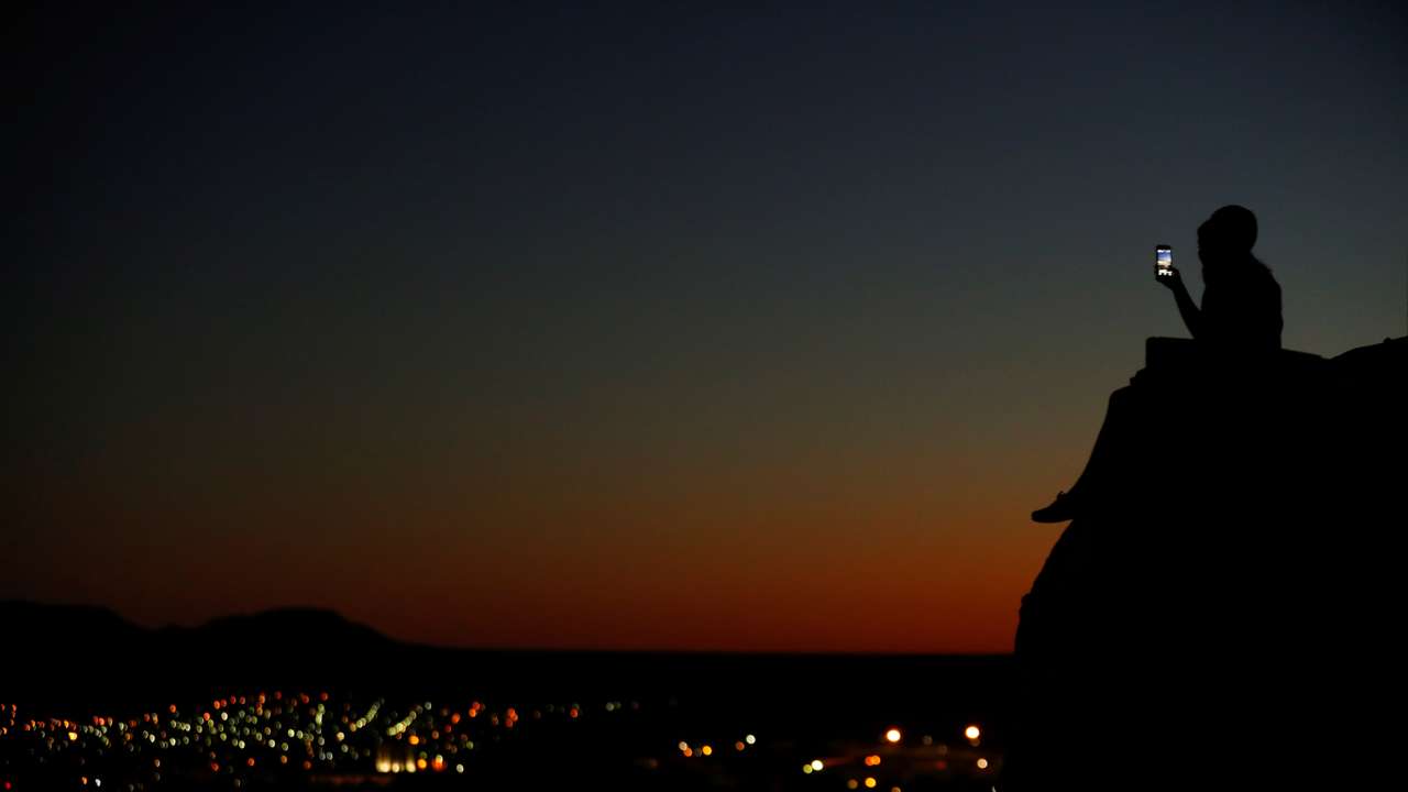 A young couple look at their phone as they sit on a hillside after sun set in El Paso, Texas
