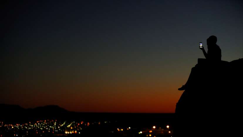 A young couple look at their phone as they sit on a hillside after sun set in El Paso, Texas